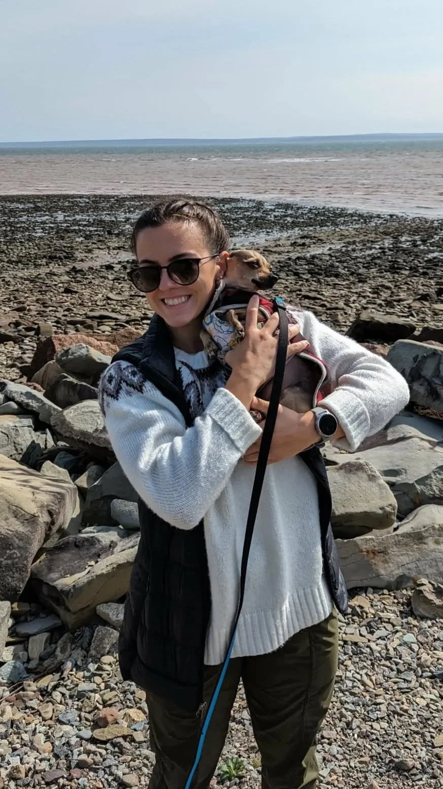 A woman smiling and wearing sunglasses, holding a small dog near a rocky shoreline with water and a cloudy sky in the background.