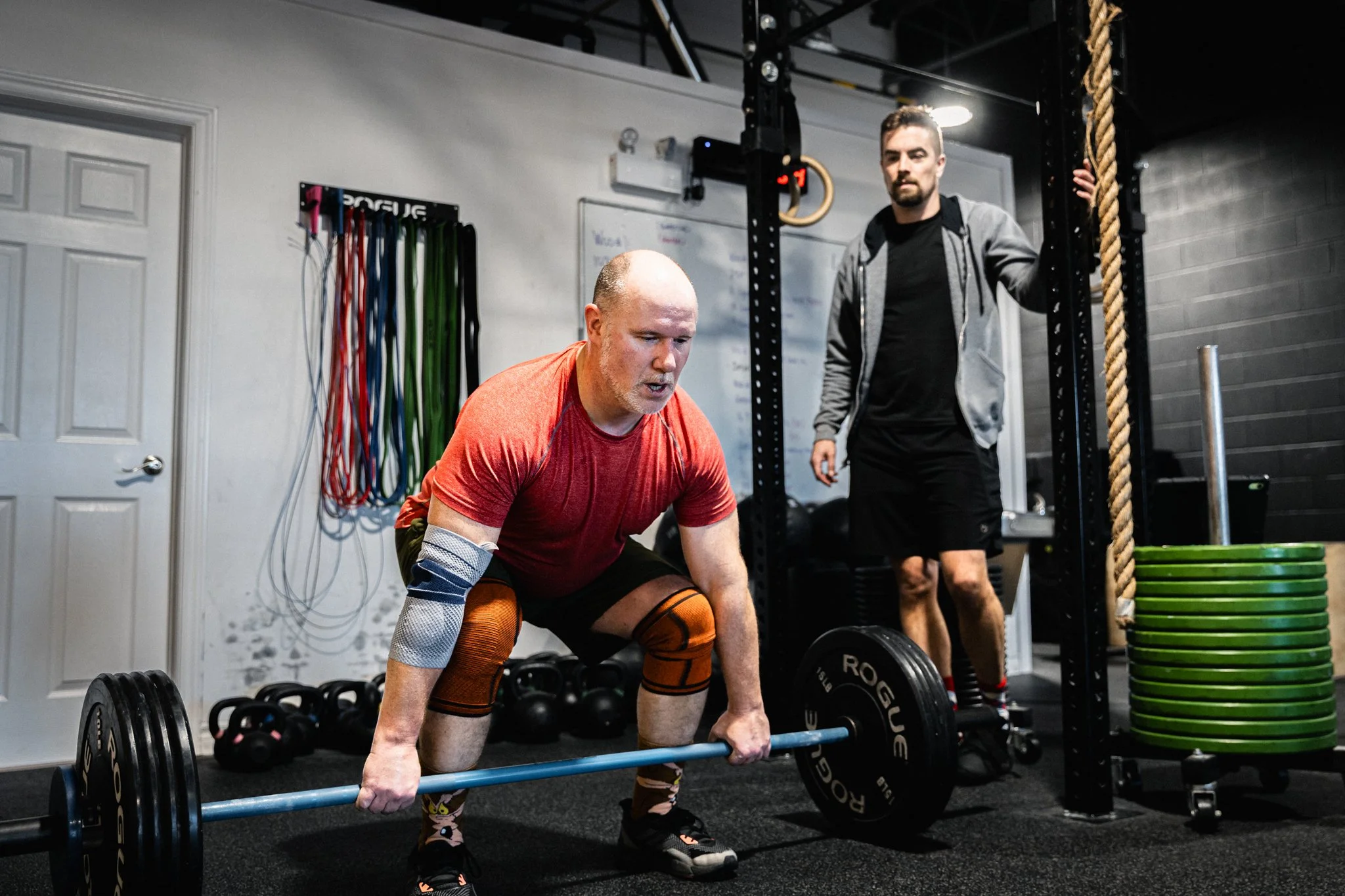 A man in a red shirt is performing a deadlift exercise with a barbell in a gym, with a trainer or friend standing nearby.