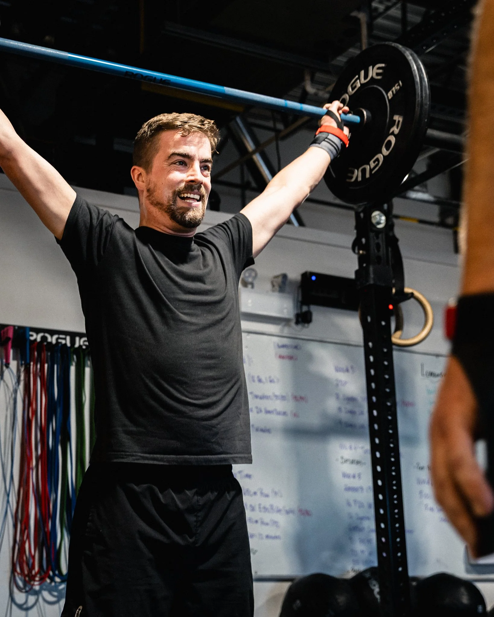 Man lifting a barbell overhead in a gym, with resistance bands hanging in the background and a whiteboard with writing on the wall.