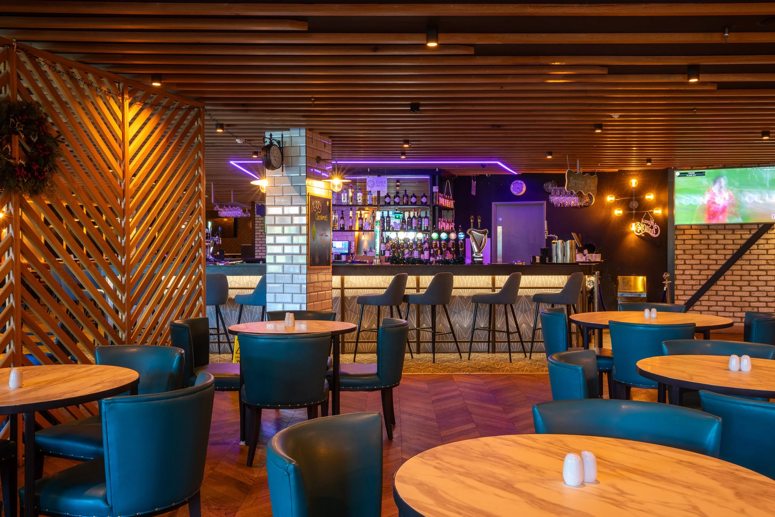 Interior of a bar with round wooden tables, blue chairs, a bar counter with bar stools, and a TV screen on the wall.