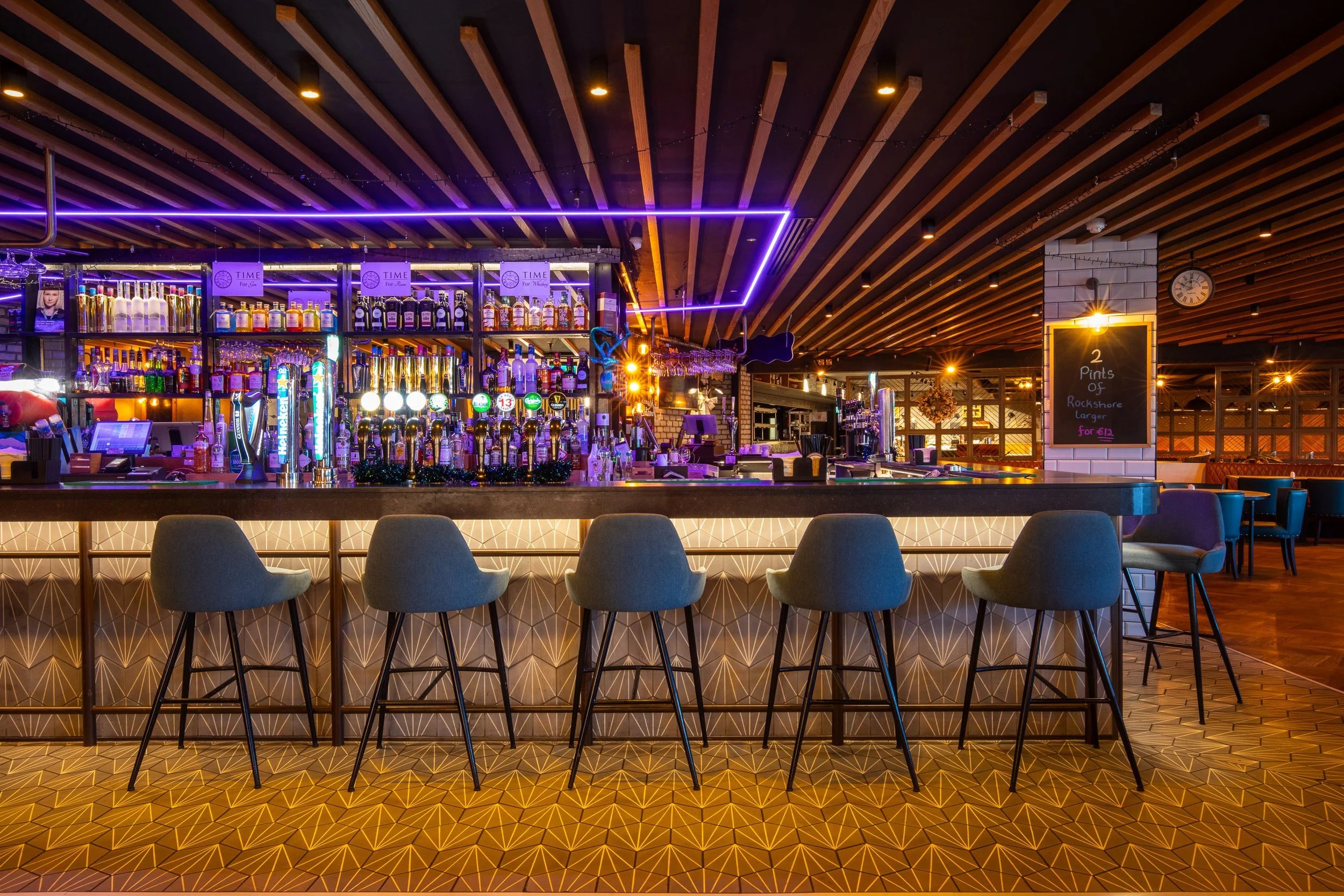 Empty bar with gray chairs in front, illuminated bottles and taps, modern lighting, and a chalkboard sign on the right.