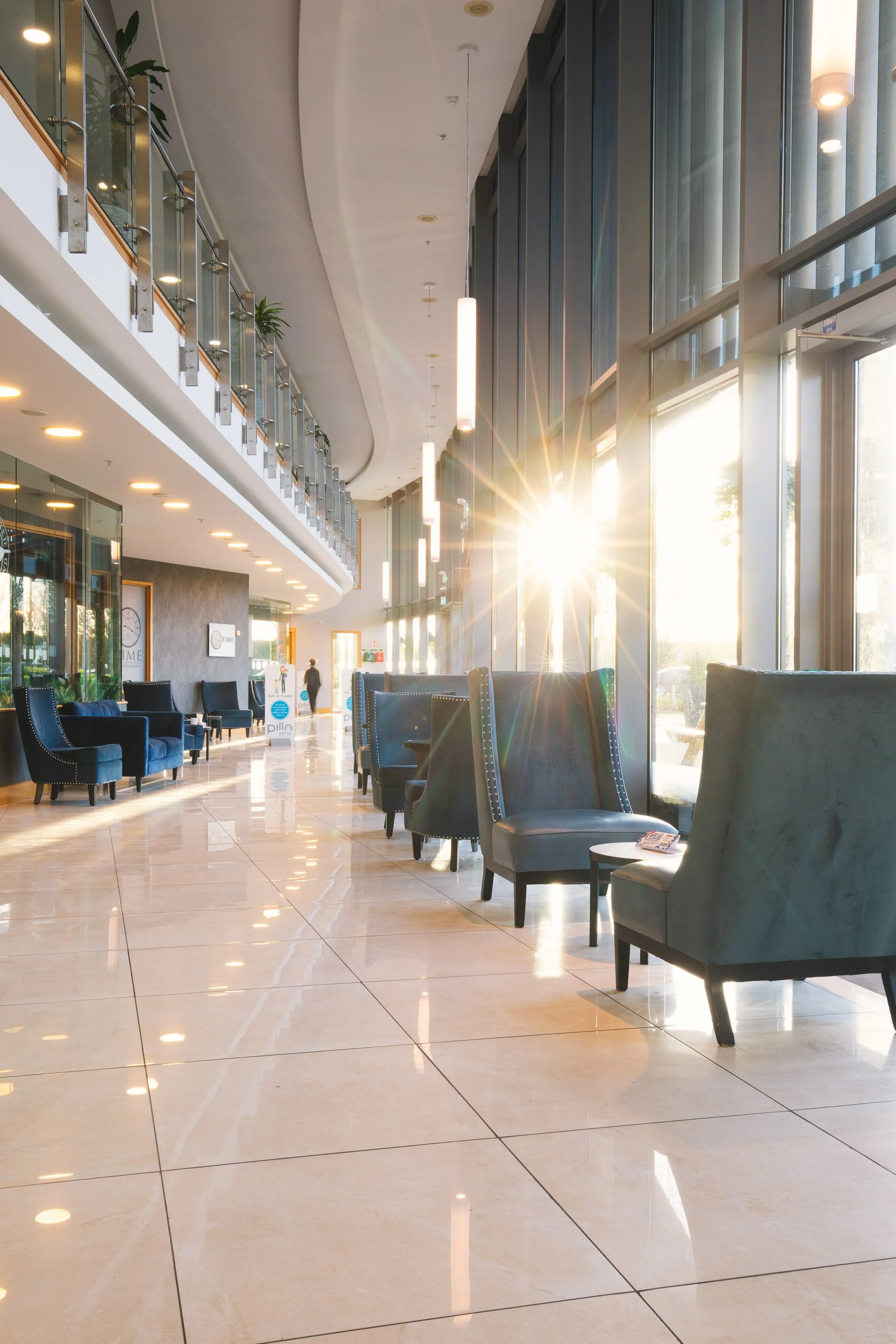 Bright hotel lobby with large floor-to-ceiling windows, blue and teal chairs, and sunlight streaming through the windows.