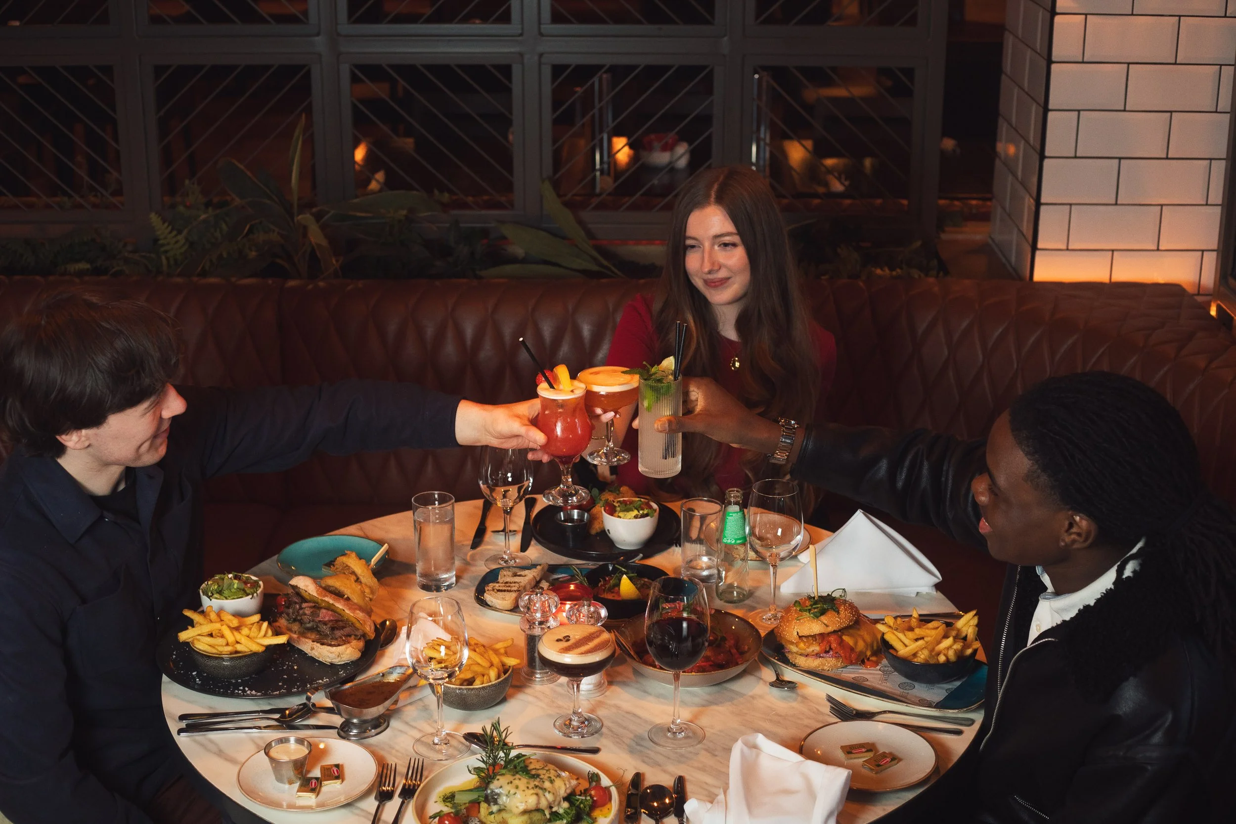 Three friends sharing drinks at a dinner table in a restaurant, toasting with colorful cocktails and smiling.