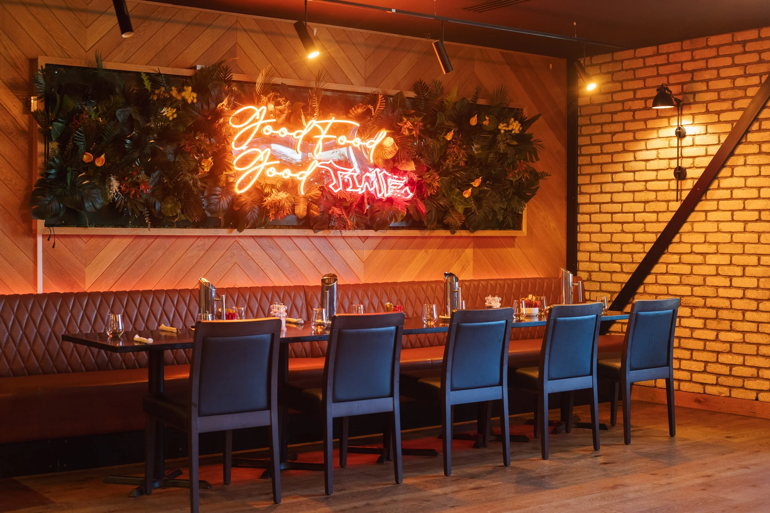 Decorative indoor restaurant setting with a long table, blue chairs, and a wall-mounted neon sign that says 'Good Food, Good Times' in cursive, with plants and wooden wall panels.