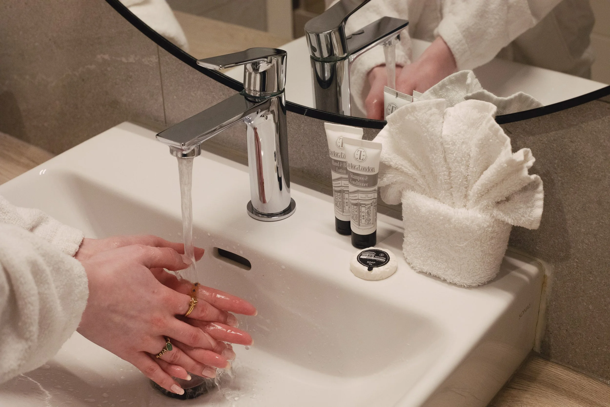 Person washing their hands at a bathroom sink with running water, a mirror, and toiletries including lotion and soap on the countertop.