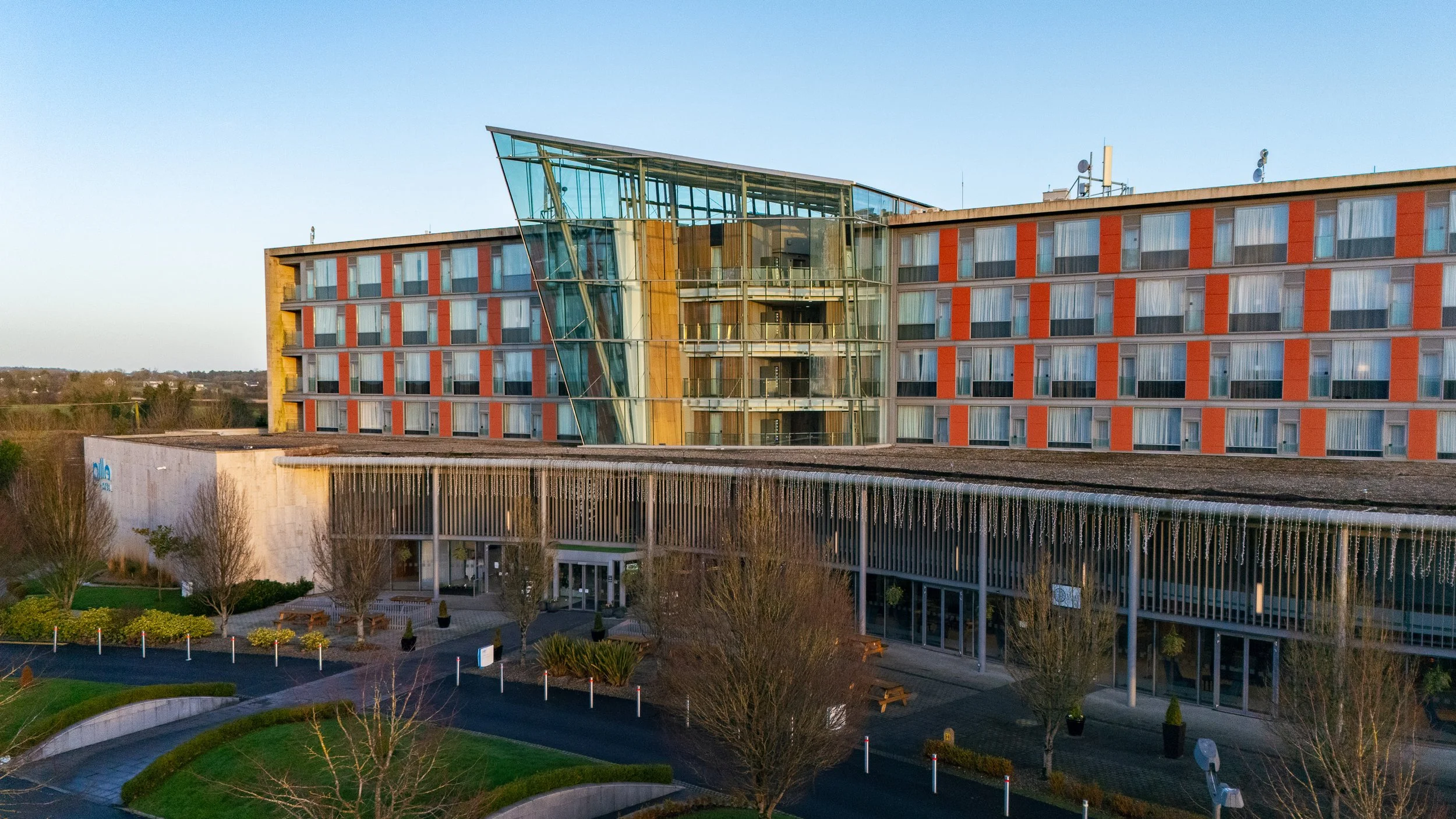 Modern multi-story hotel building with a glass front and orange accents, surrounded by landscaped outdoor area with trees and benches.