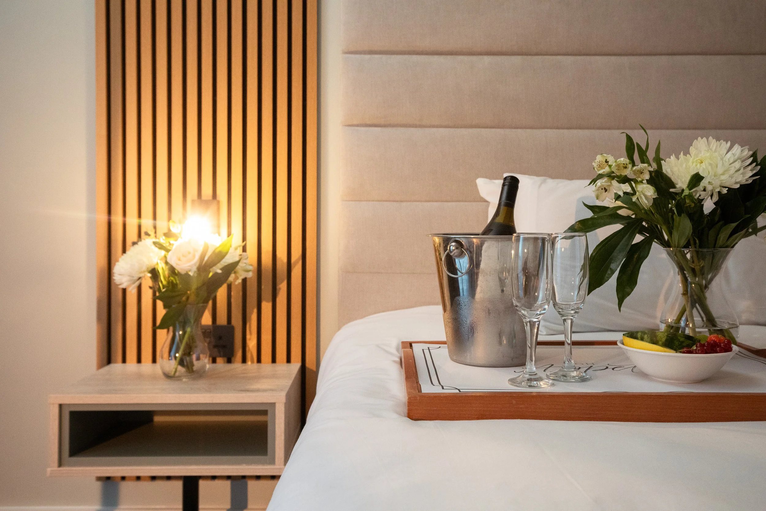 Bedside table with a vase of white flowers, a tray with a wine bottle in an ice bucket, two empty champagne glasses, and a bowl of fresh fruit in a hotel room.