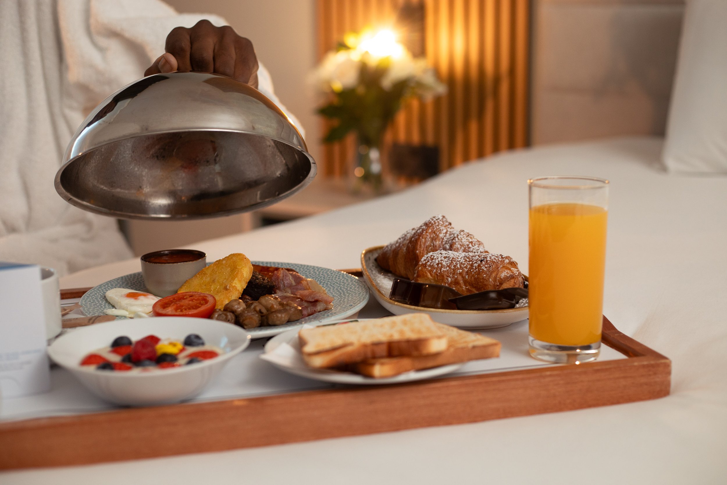 Breakfast tray with toast, croissants, fruit, yogurt with berries, bacon, eggs, tomato, mushrooms, and orange juice on a bed in a cozy room with warm lighting.