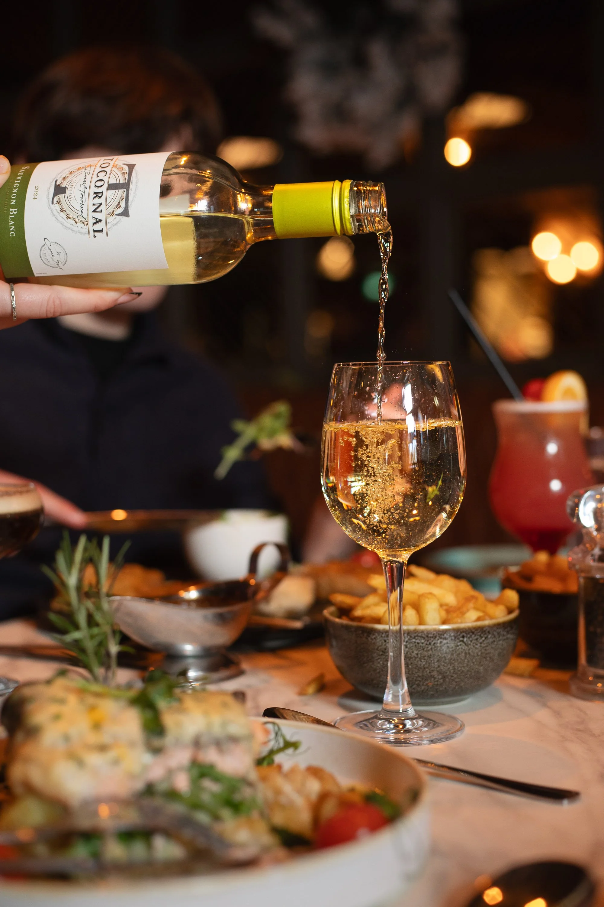A hand pouring white wine into a glass at a dinner table with food and drinks, in a warmly lit restaurant setting.