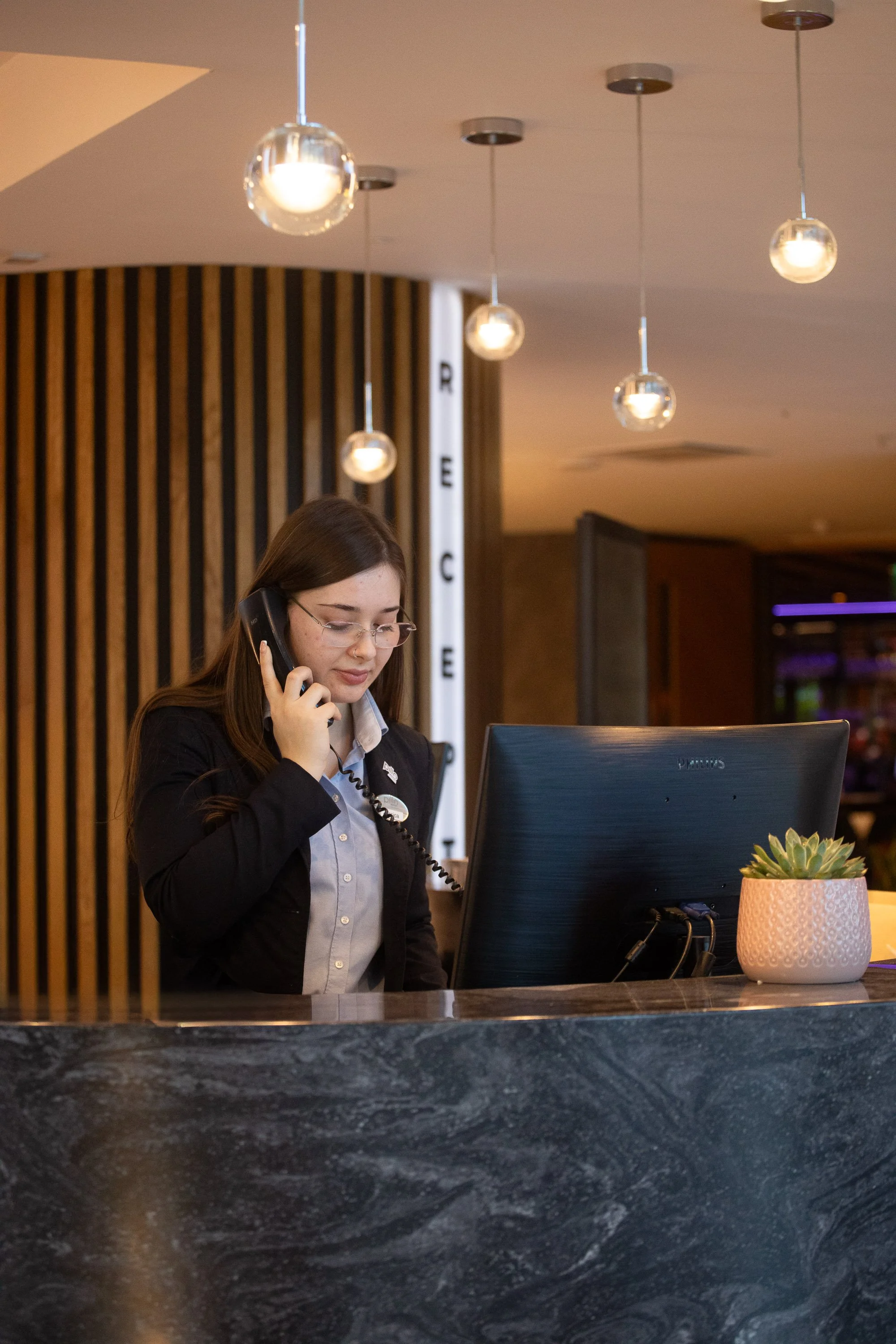 A woman with long brown hair, glasses, and a black blazer talking on a landline phone at a hotel reception desk, with a computer monitor and a potted succulent plant on the desk.