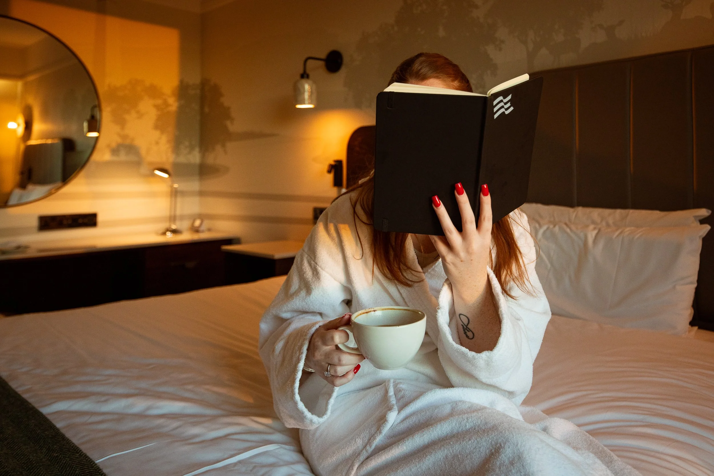 A woman with red hair and red nail polish sitting on a bed, holding a cup of coffee or tea in one hand while covering her face with a book in a cozy, warmly lit bedroom.