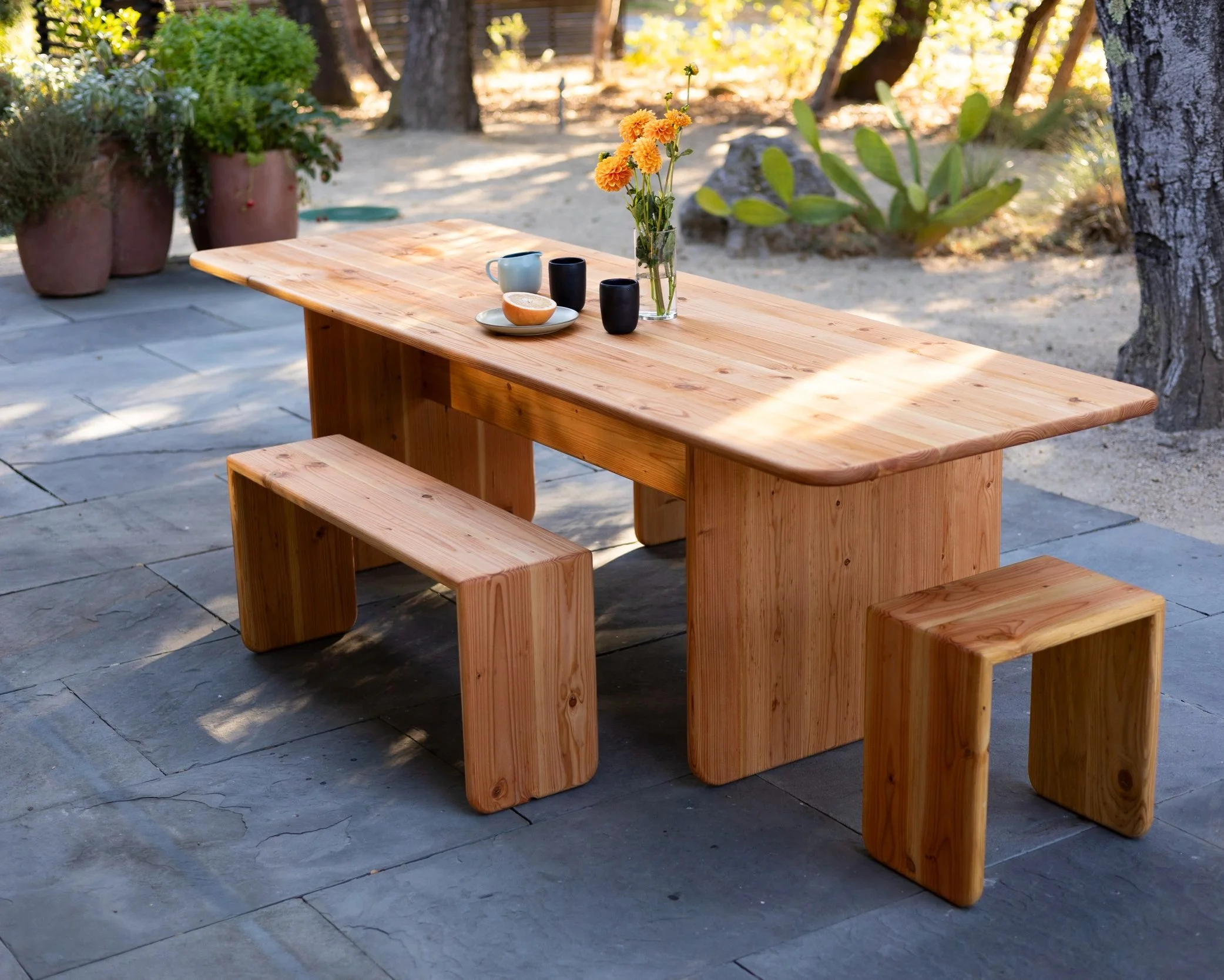A wooden outdoor dining table with matching benches on a stone patio, with potted plants and trees in the background.