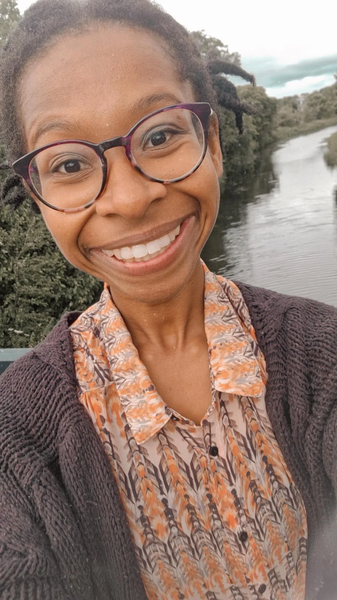 Natasha wearing glasses taking a selfie near a river with trees in the background.