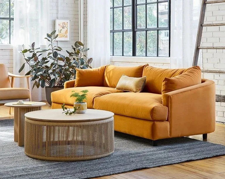 A modern living room with a mustard yellow sofa, a light beige armchair, a round coffee table, and large windows with white curtains, decorated with a potted plant and framed artwork.