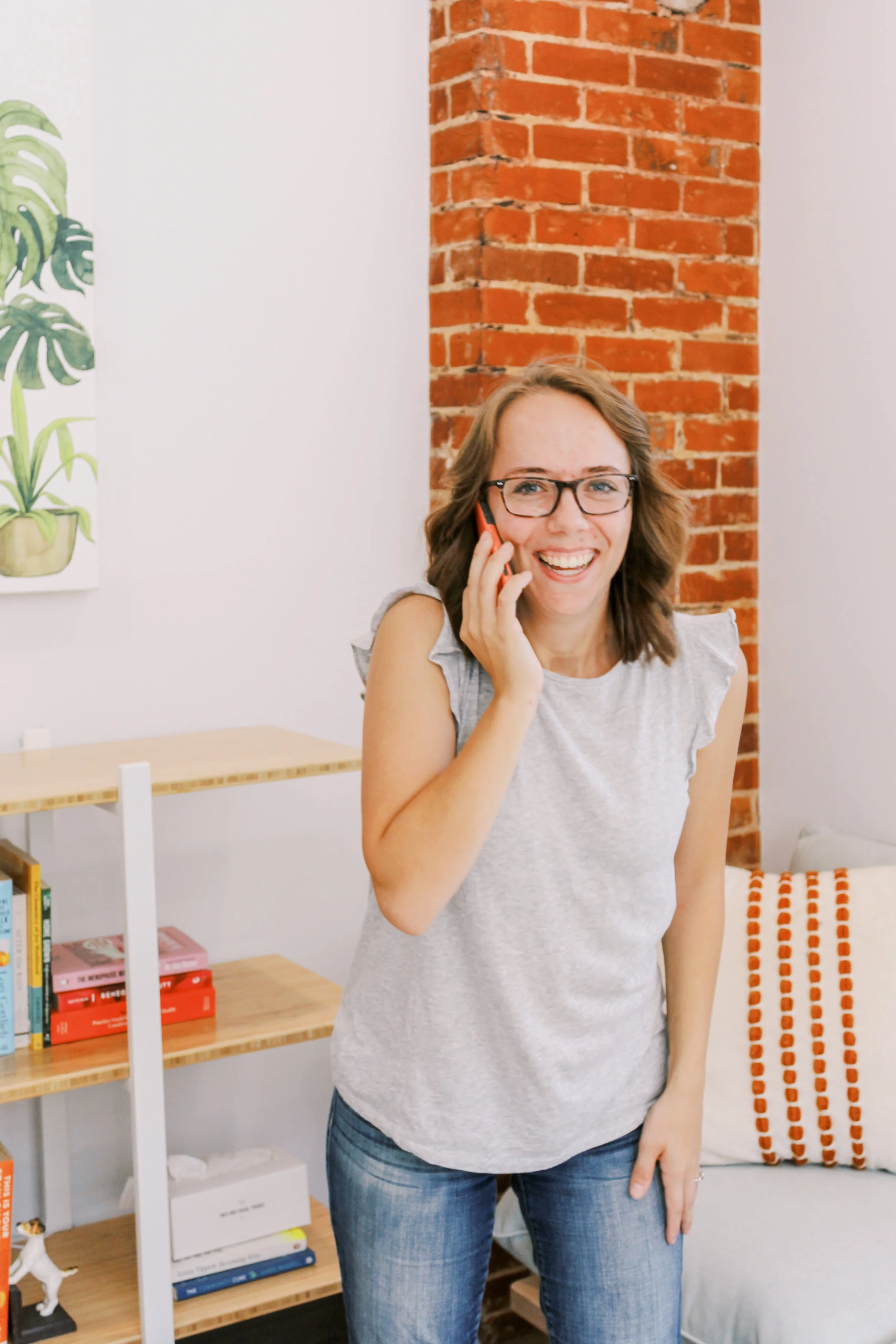 A woman with glasses smiling while talking on her cellphone in a modern, cozy room with a brick accent wall, a colorful pillow, and a bookshelf.