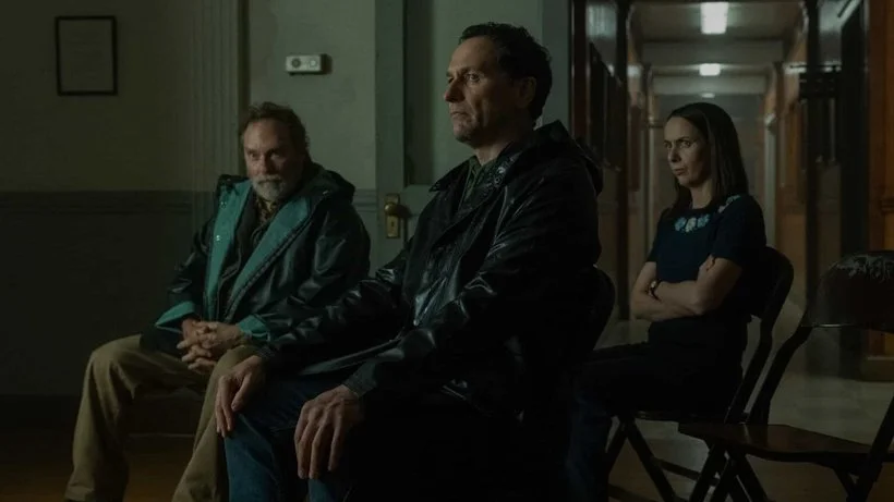 Wyck (Stephen Root), Tom (Matthew Rhys), and Patricia (Kate O'Flynn) seated in folding chairs in Widow’s Bay Town Hall.