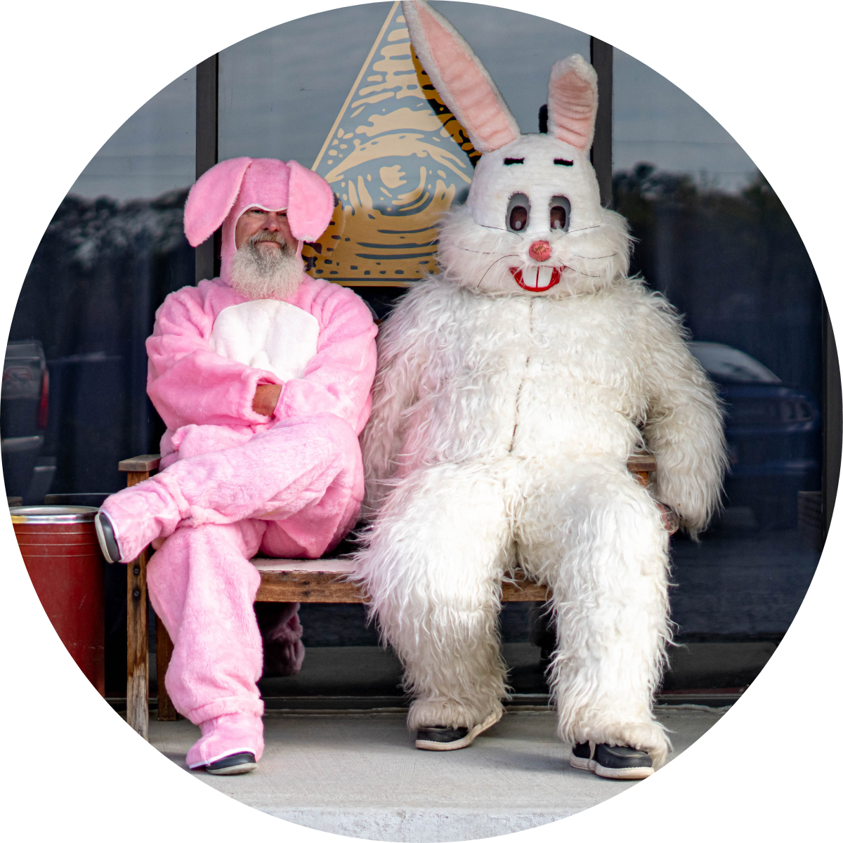 Two people dressed in elaborate animal costumes, one as a pink bunny and the other as a white furry rabbit, sitting on a wooden bench in front of a window with a gold geometric art piece.