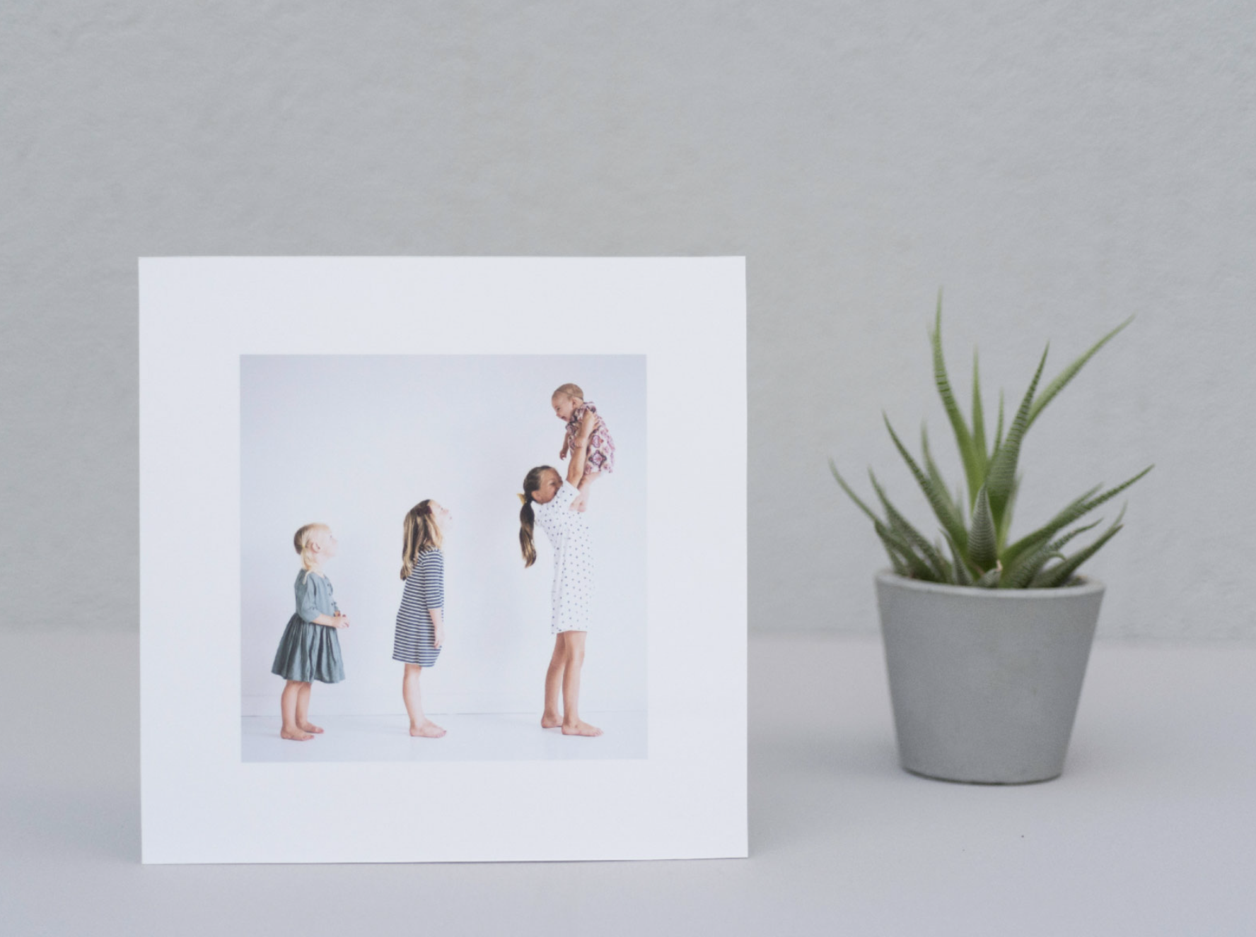 A family photograph displayed in a white frame, featuring a woman holding a baby, with two children beside her. A small potted succulent is next to the frame on a gray surface.