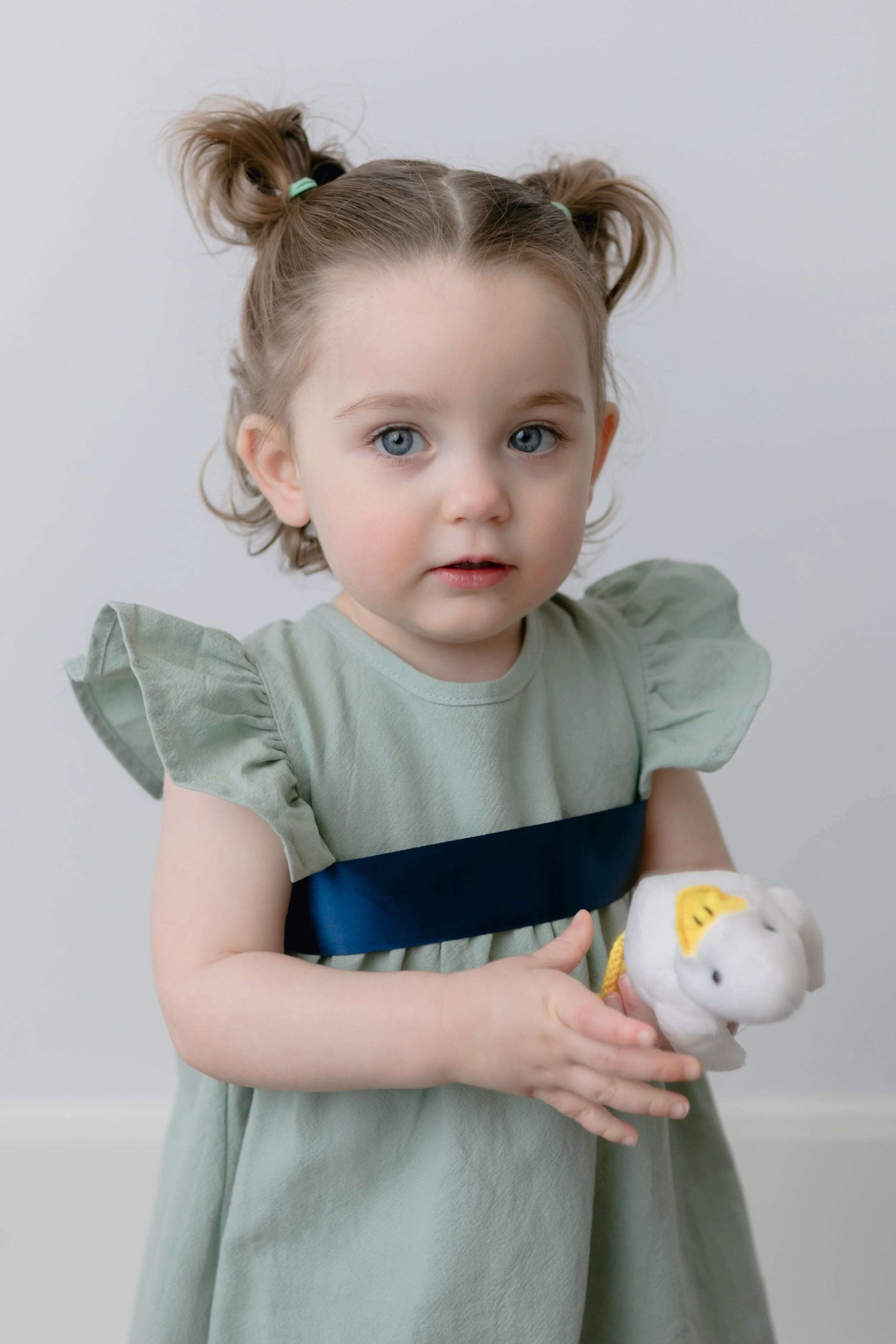 Toddler with blue eyes wearing green dress holds a small stuffed toy.