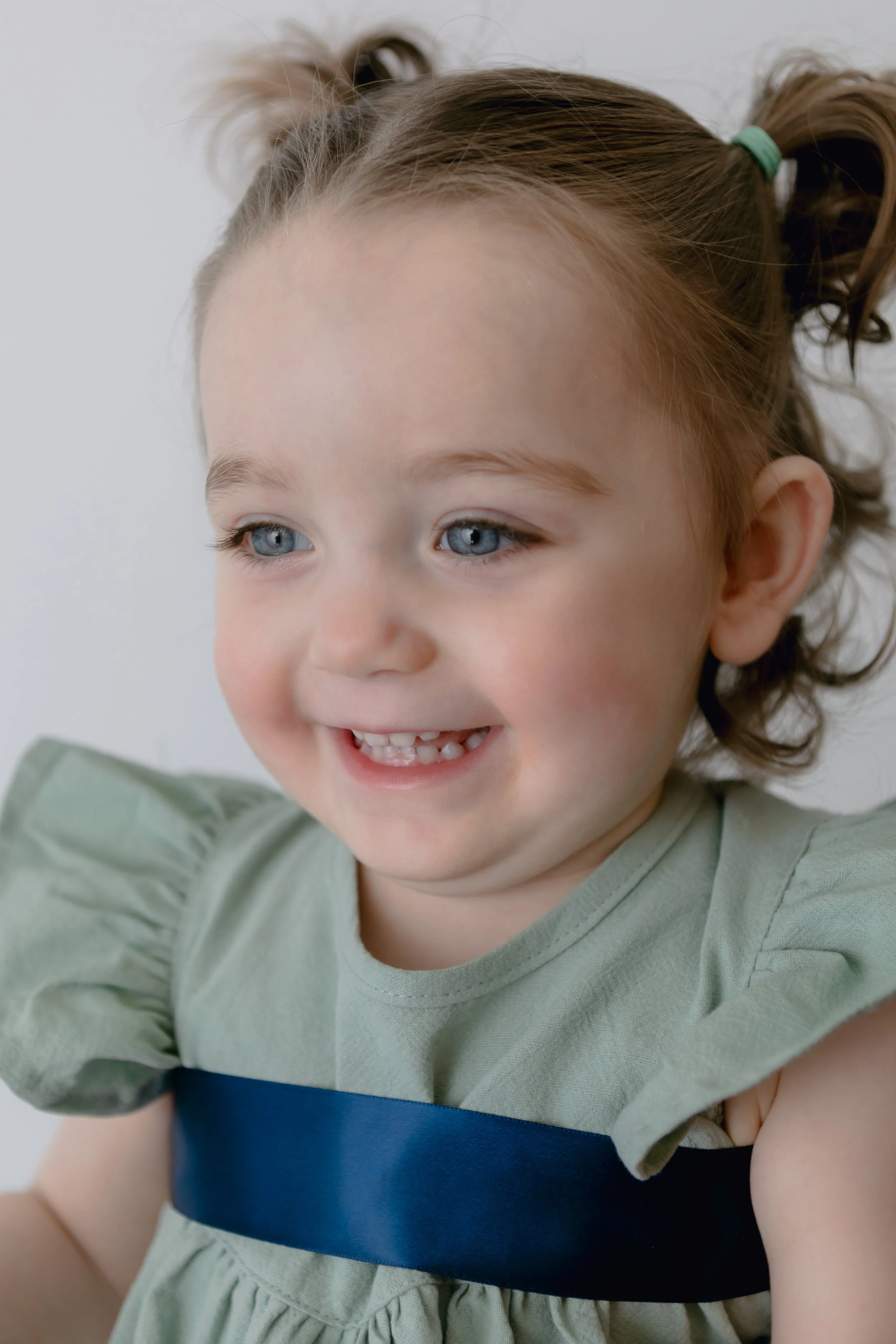 Smiling toddler with light blue eyes wearing a light green dress with a navy blue band, hair in pigtails.