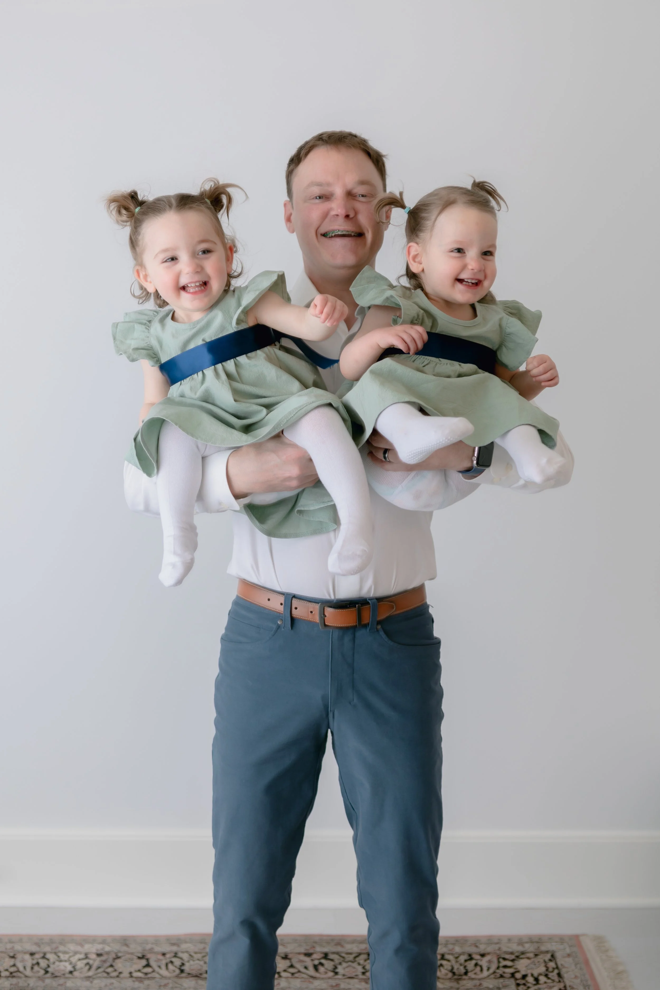 A man smiling and holding two young girls, each wearing matching light green dresses with blue sashes and white leggings. The man is wearing a white shirt and blue pants with a brown belt against a plain white background.