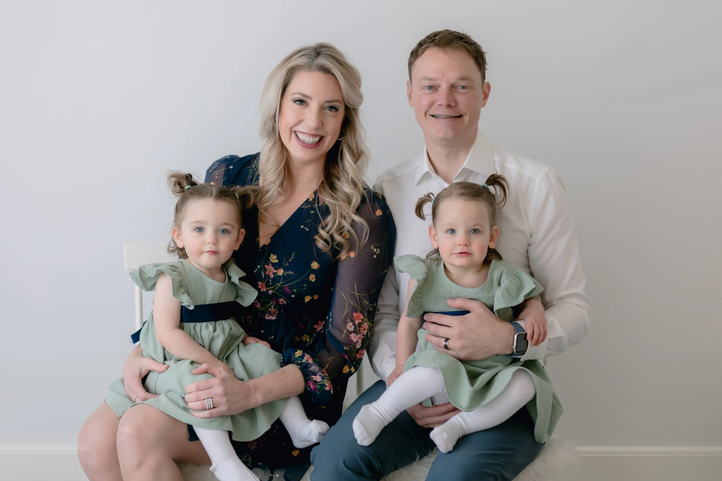 Smiling family with two young children wearing light green dresses, posing against a neutral background.