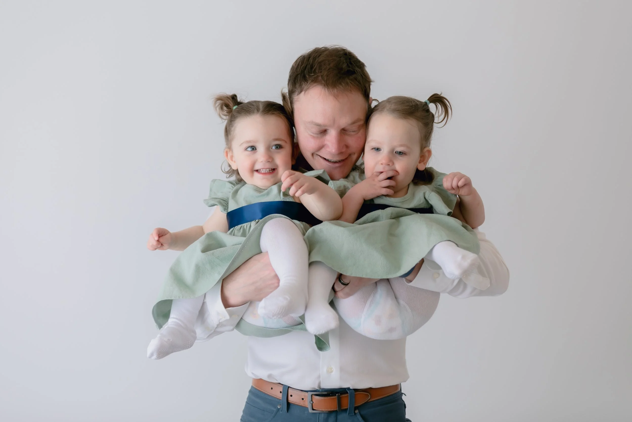 A person holds two young children, each wearing matching light green dresses with navy blue sashes. The children have small pigtails and are smiling. The person is wearing a white shirt and gray pants.