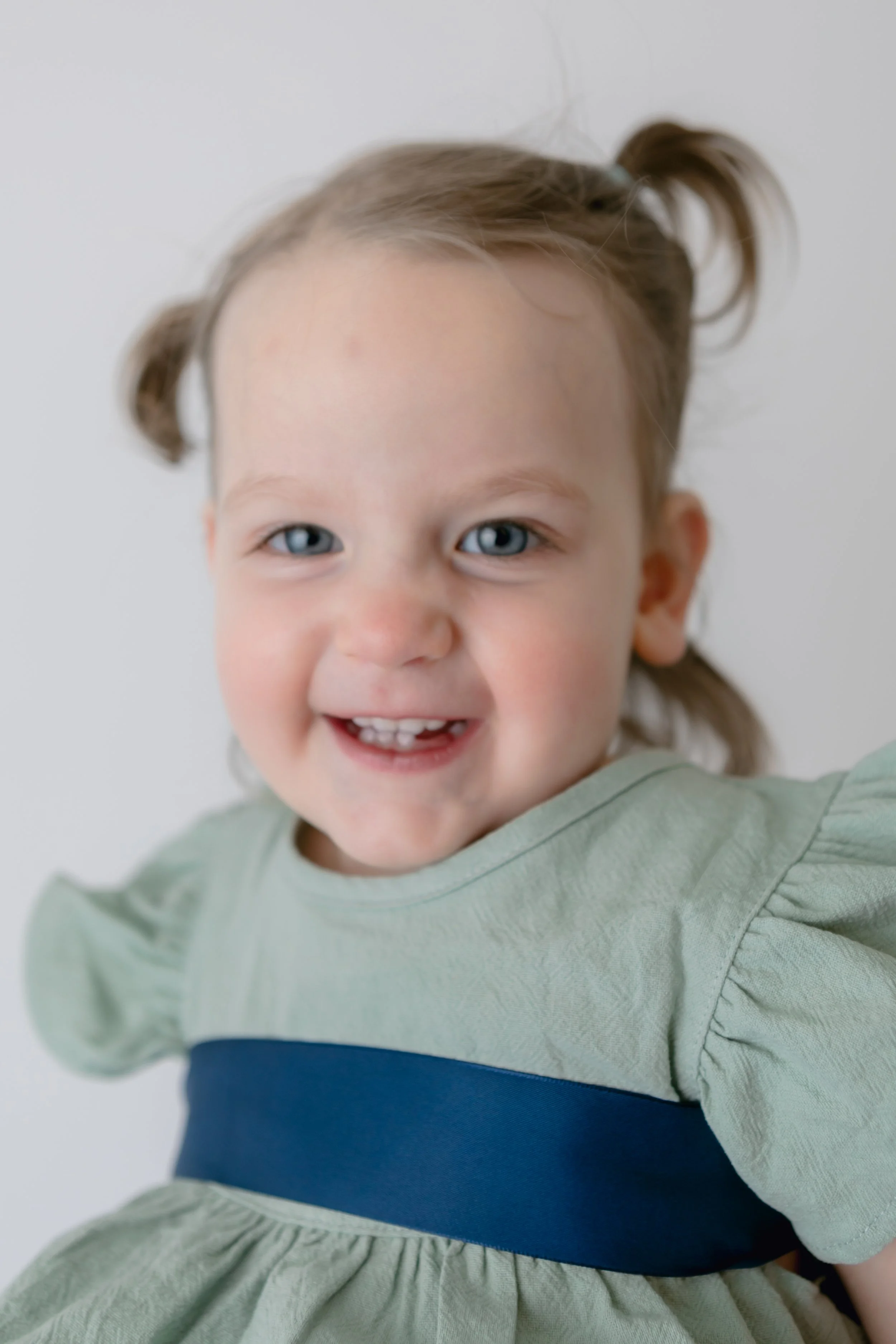 Smiling toddler with light hair in pigtails wearing a green dress with blue band