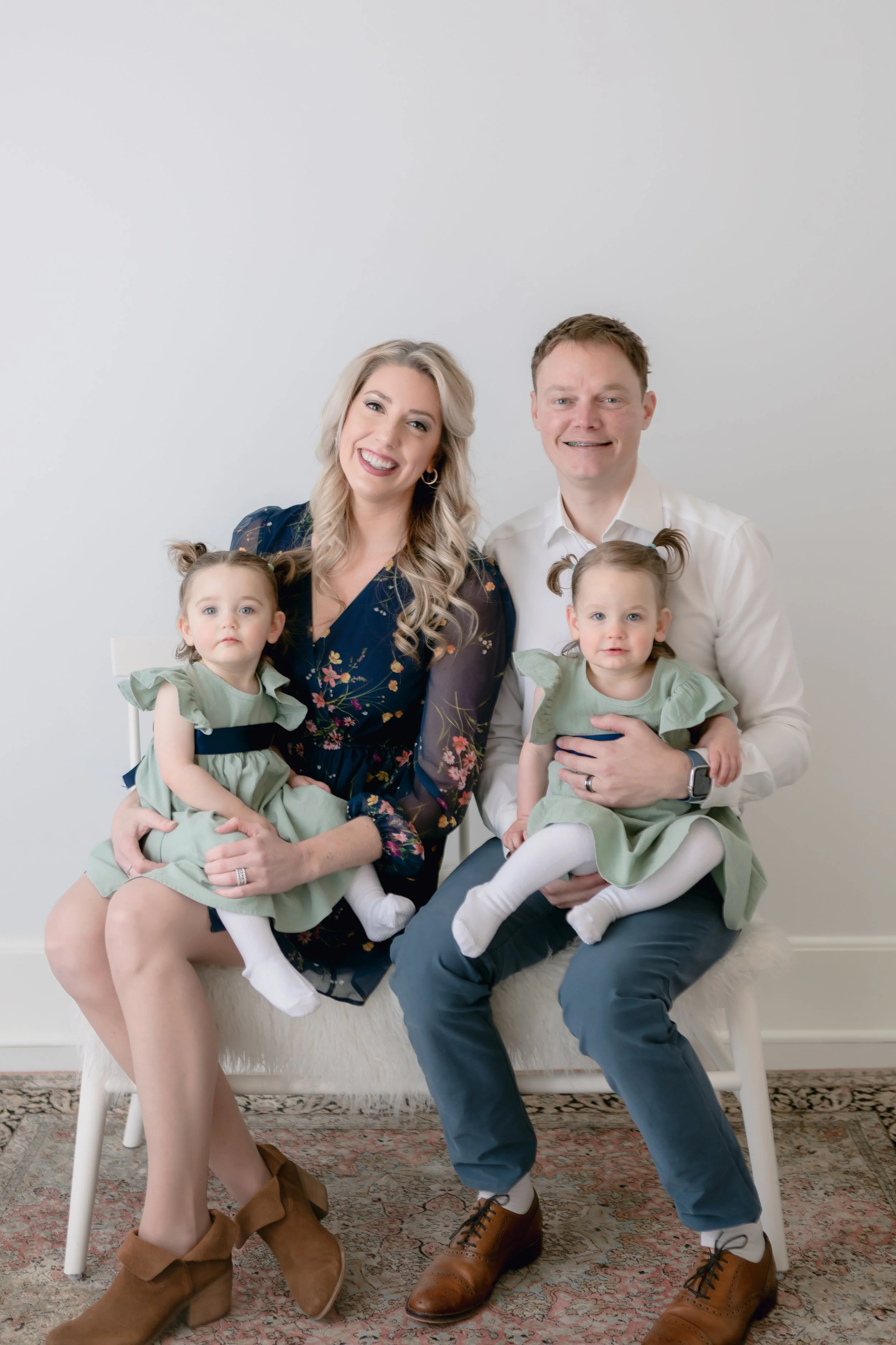 A family portrait featuring two adults and two young children sitting on chairs against a white wall, with the children wearing matching green dresses and the adults dressed in formal attire.