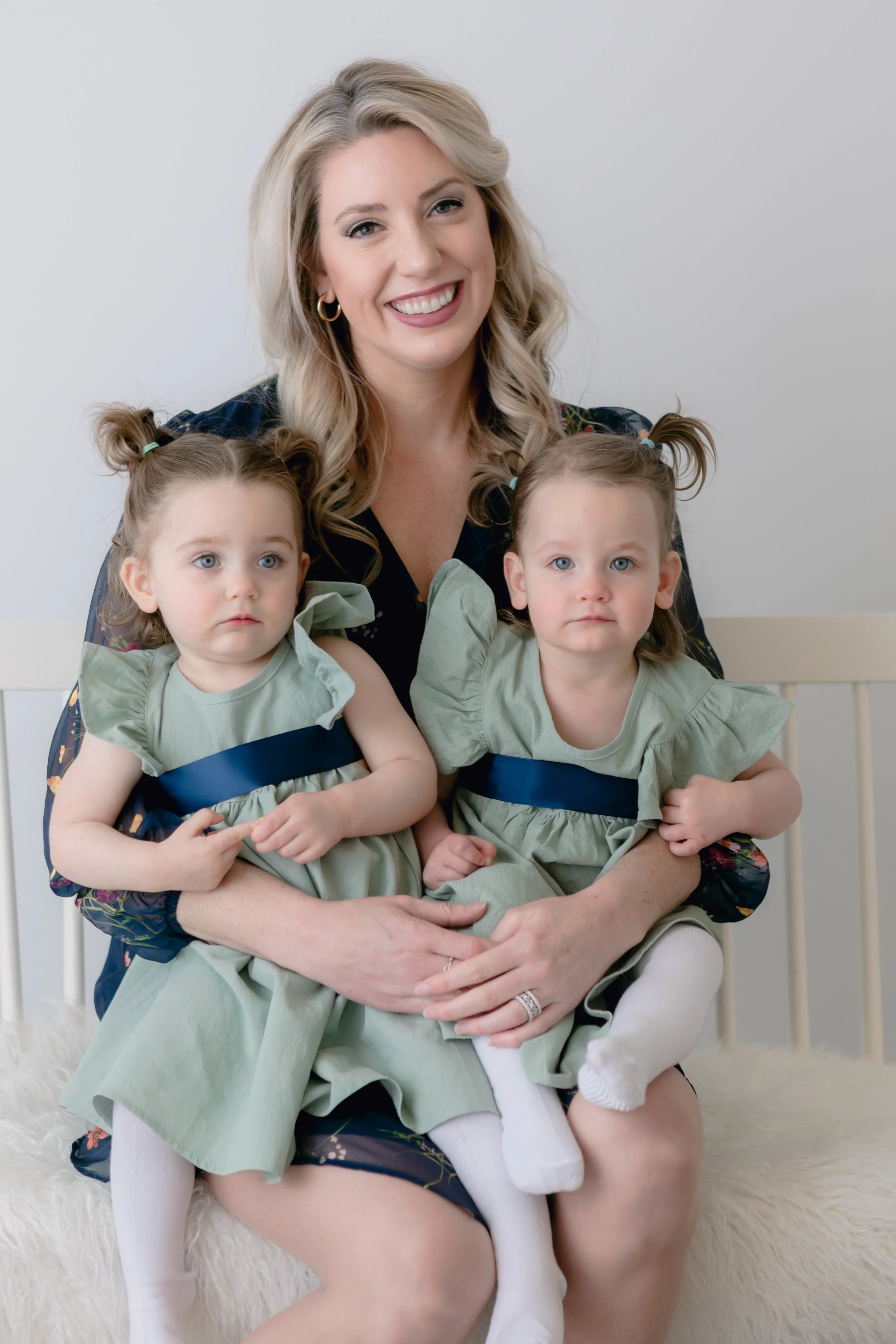Woman smiling while holding twin toddlers wearing matching green dresses with navy blue sashes.