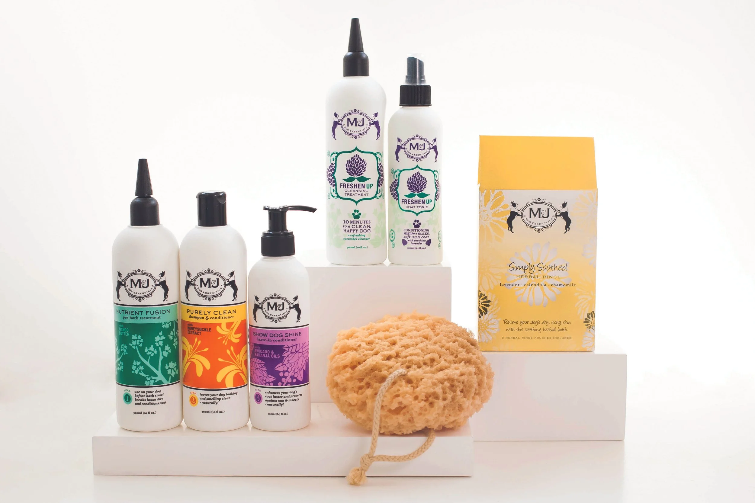 A collection of MJ dog grooming products including shampoo, conditioner, pre-bath treatment, coat tonic, and a herbal rinse. A natural sponge is also displayed beside the products.