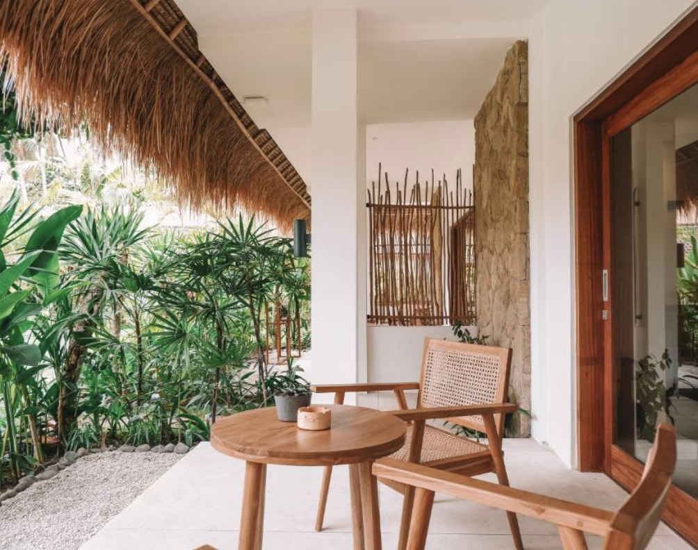 Outdoor patio with wooden furniture and lush greenery, featuring a round table, a chair with woven backrest, and a thatched roof overhead.