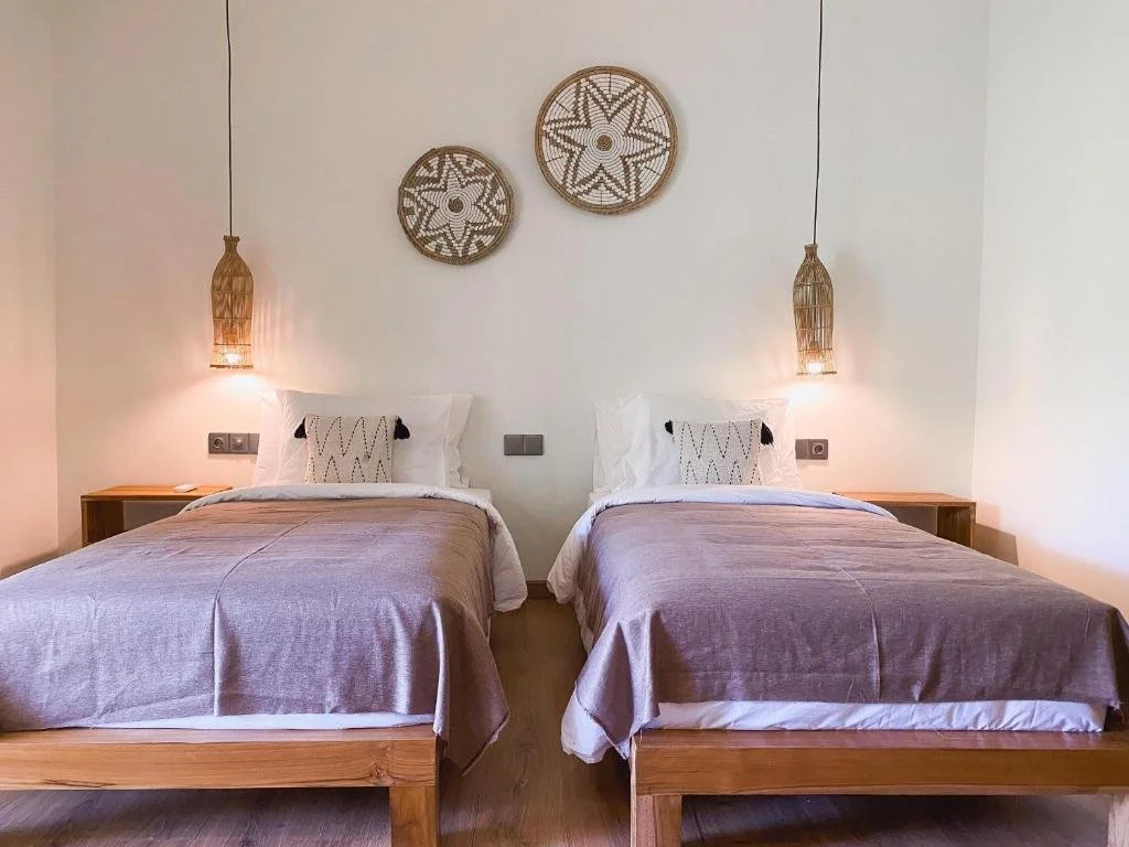 A hotel room with two beds, each with white pillows and beige blankets, a wooden nightstand, and woven hanging lamps. There are two wall decorations above the beds with star patterns.