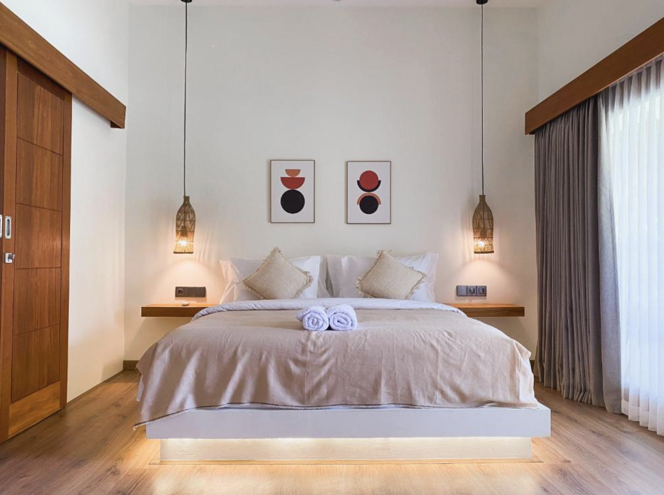 A neatly made bed in a modern bedroom with beige bedding and pillows, two rolled white towels, hanging wicker pendant lights, wooden floating side tables, abstract art prints on the wall, and curtains on a window to the right.
