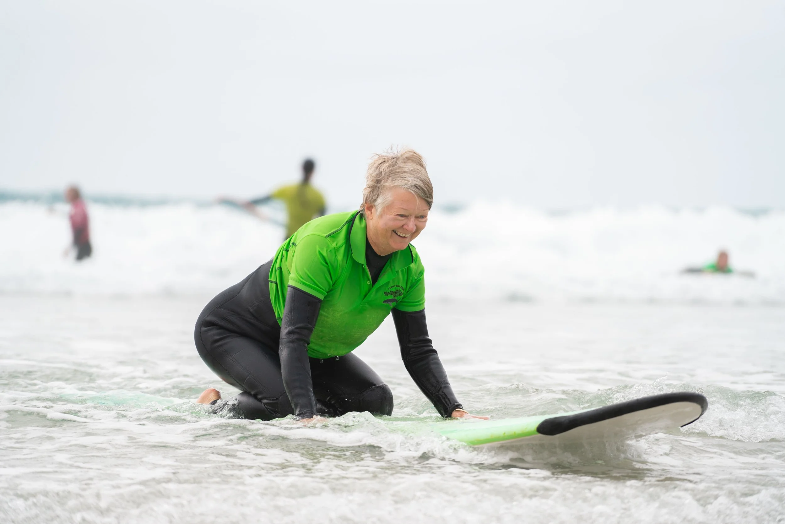 Older woman in a bright green shirt and black wetsuit kneeling in shallow ocean water, smiling, with a surfboard in front of her and other people in the background