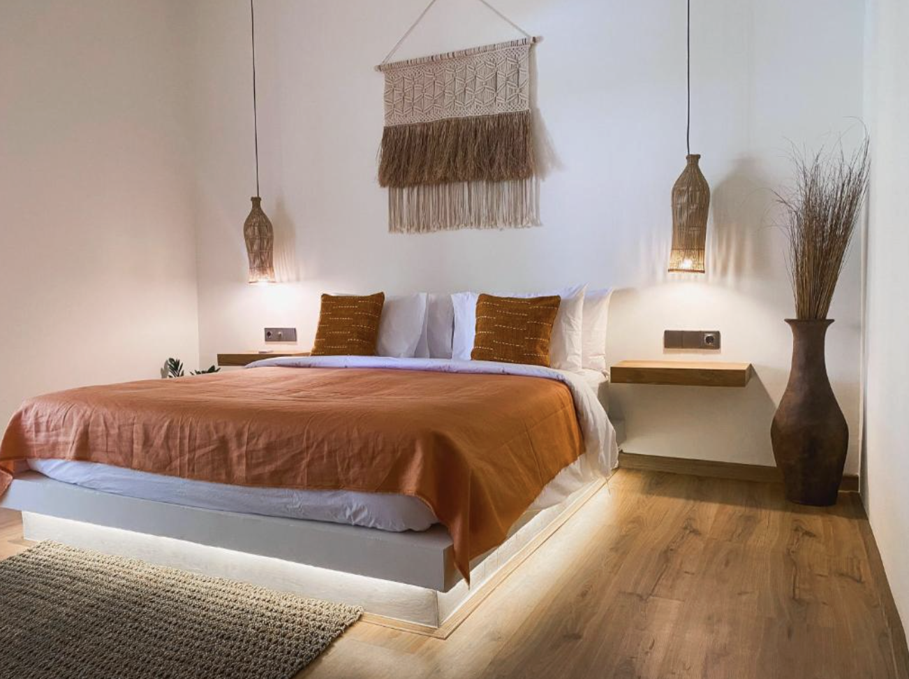 A cozy bedroom with a bed that has white and rust-colored bedding and pillows. Two hanging pendant lights, a woven wall hanging, and a large vase with dried branches are on the wall and beside the bed, with soft lighting creating a warm atmosphere.