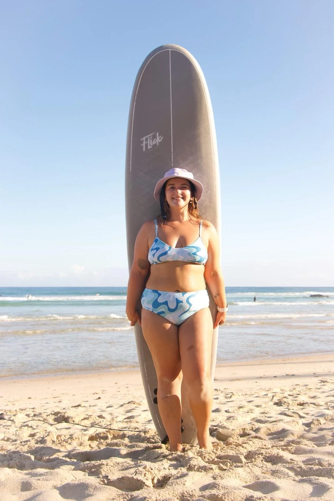 A woman in a patterned blue and white swimsuit standing on a sandy beach, holding a surfboard behind her, with the ocean in the background and a clear blue sky.