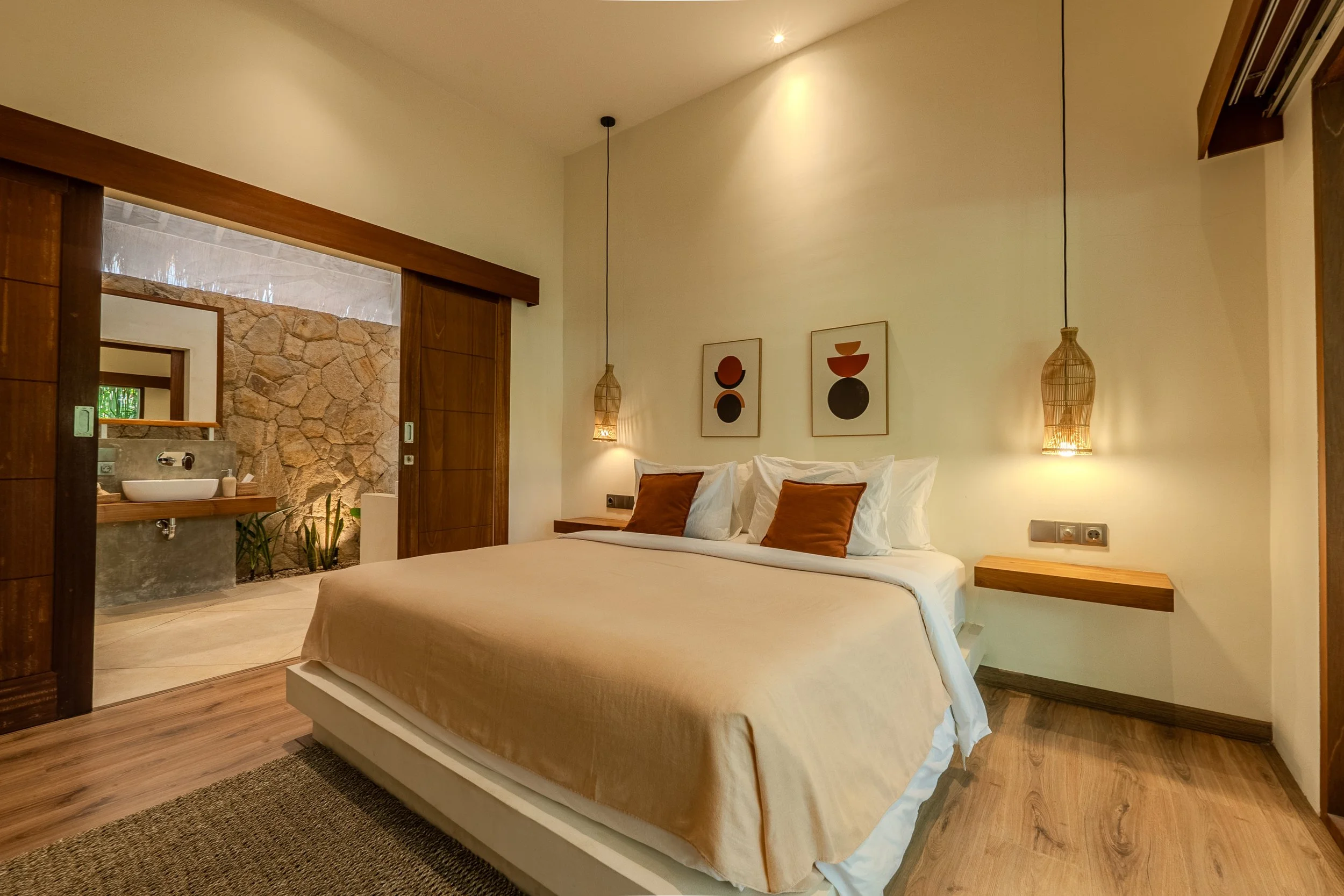 A cozy bedroom with a large bed, white linens, and brown accent pillows, two hanging pendant lights, and artwork above the bed, with an open doorway leading to a stone-walled bathroom.