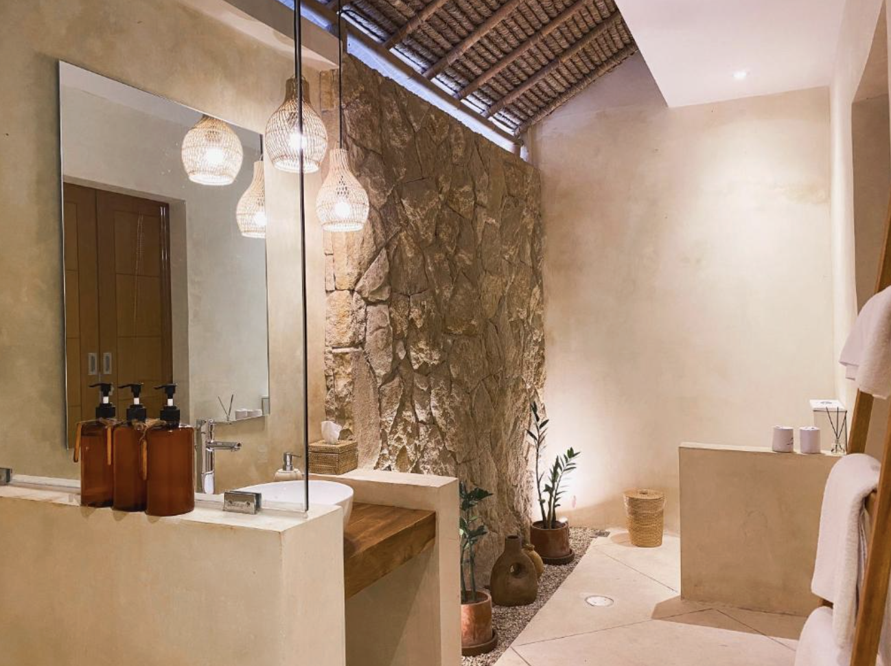 Stylish bathroom with stone accent wall, hanging wicker lamps, a large mirror, bottles of soap, potted plants, and neutral-colored decor.