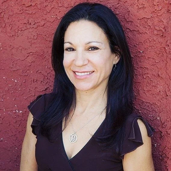 Portrait of a woman with long dark hair, wearing a dark purple top and a silver necklace, standing in front of a textured red wall.