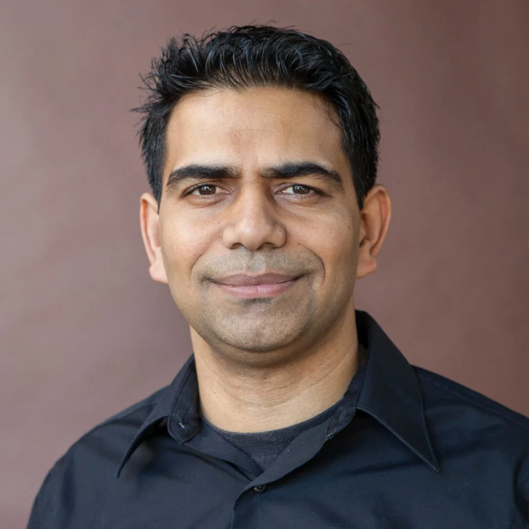 A man with black hair and brown eyes, wearing a black collared shirt, smiling gently, standing against a plain brown background.