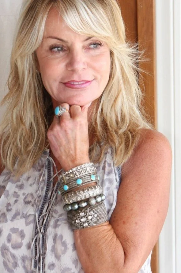 A woman with blonde hair wearing multiple silver and turquoise bracelets, a ring, and a patterned top, posing indoors with a thoughtful expression.