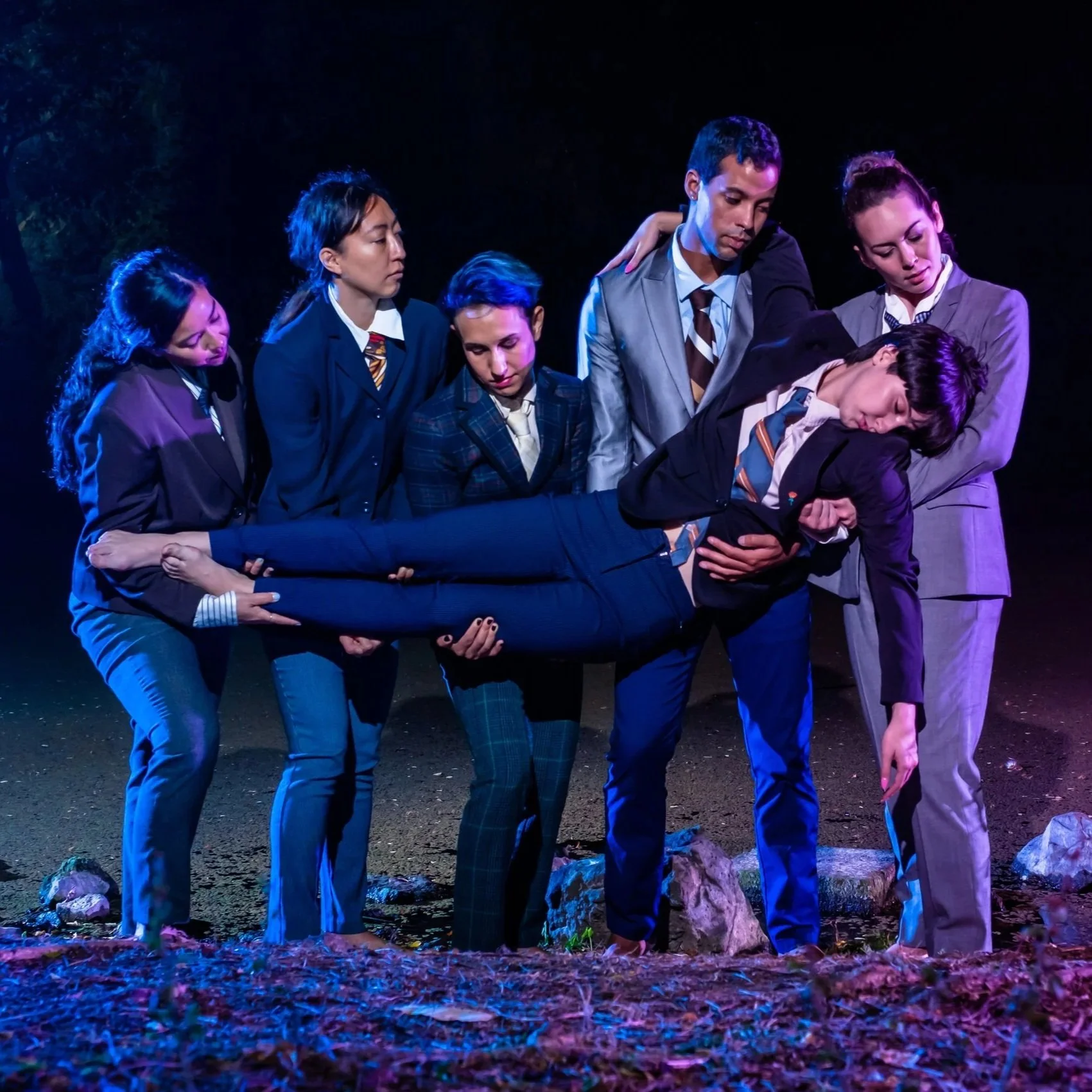 The cast of "Never Forever," a surrealist contemporary dance theatre production choreographed and directed by Maria Teresa Houar, appear in front of a lake wearing suits, and carry one dancer who appears to be sleeping.