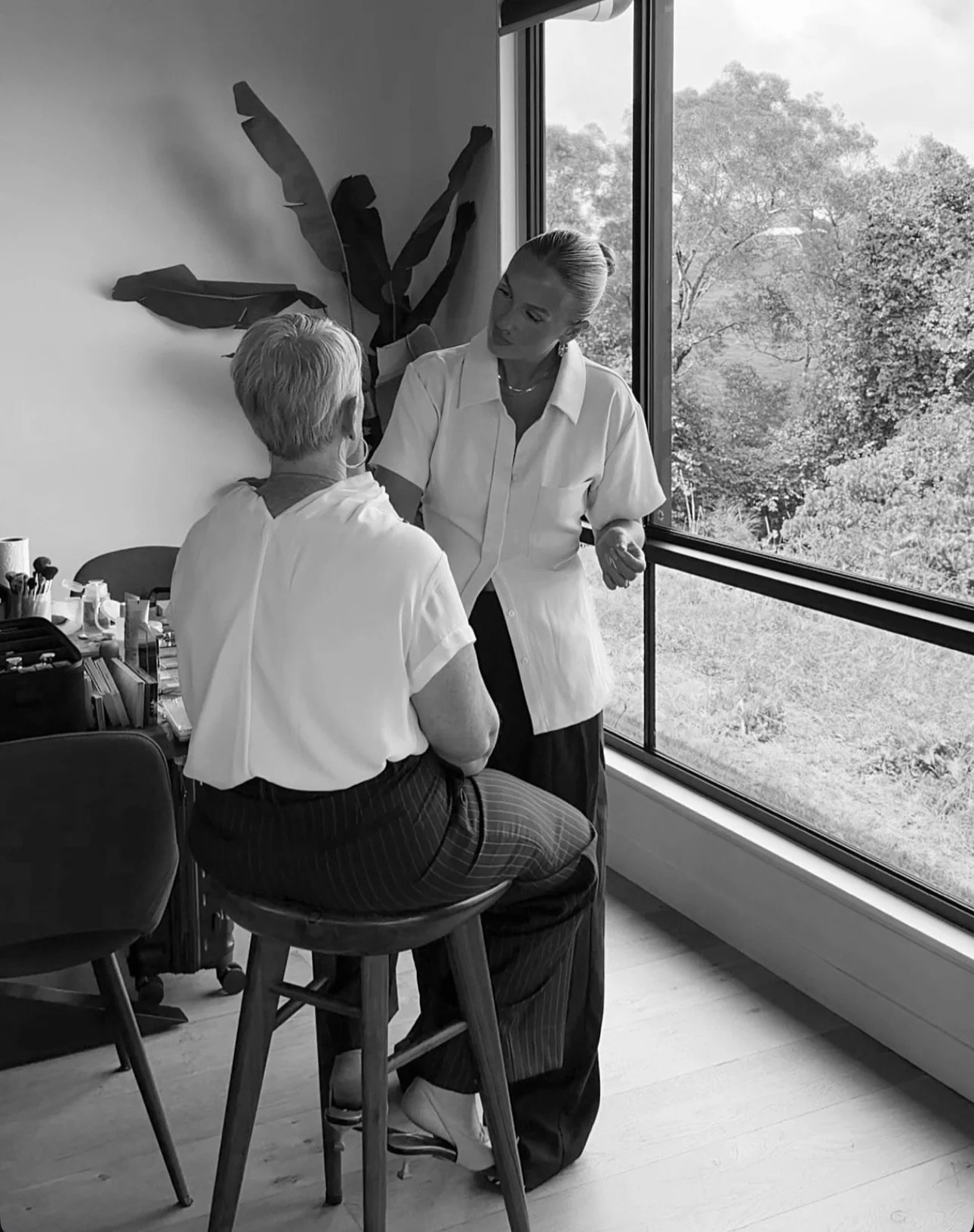 A woman healthcare professional talking to an elderly patient in a room with large windows showing trees outside.