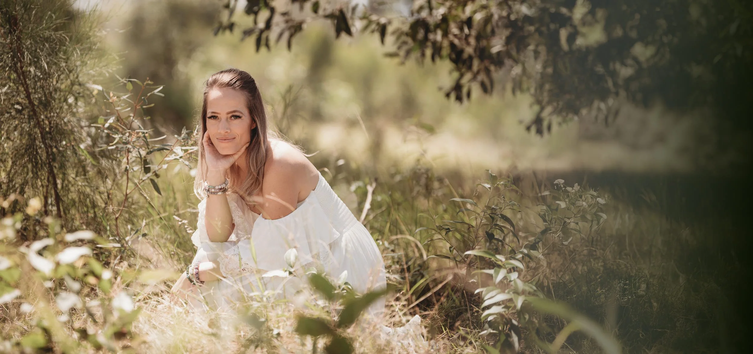 Cas crouches in a field with long grass and trees, wearing a white dress and crystal bracelets