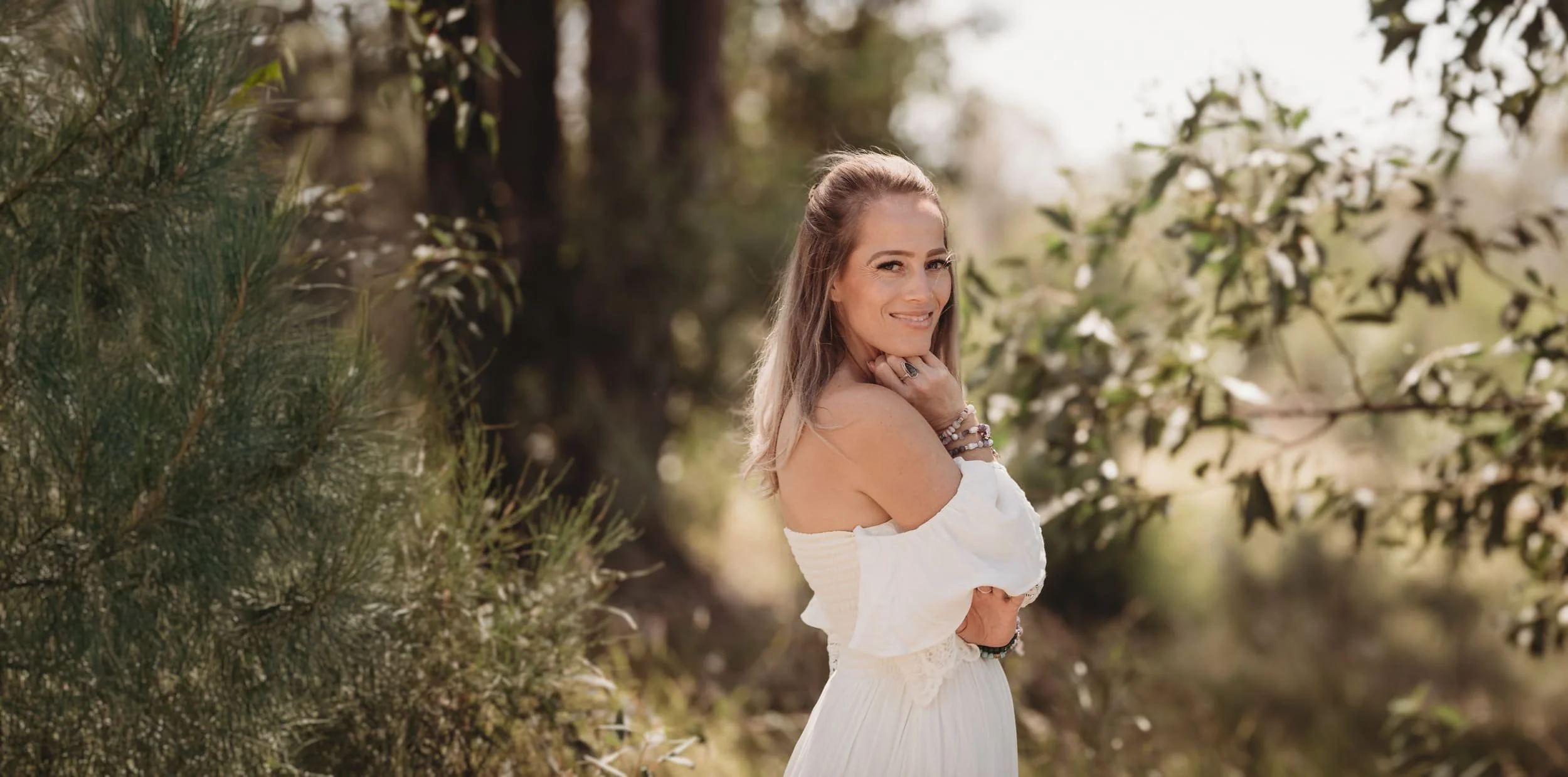 Cas, human design practitioner, stands in a forest wearing a white dress smiling side on over her shoulder at the camera