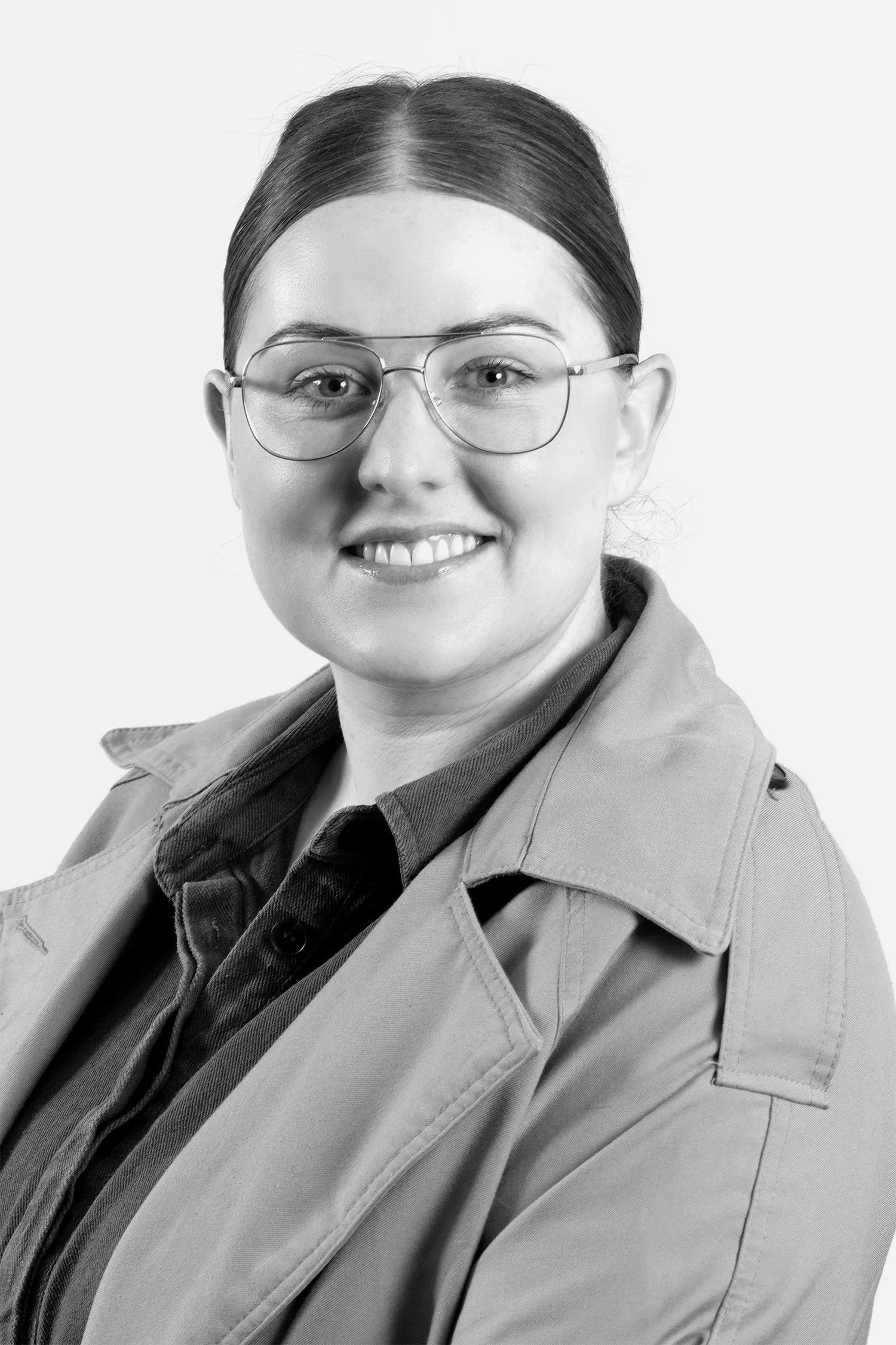 Black and white photo of a woman smiling, wearing glasses, a dark shirt, and a light-colored jacket.