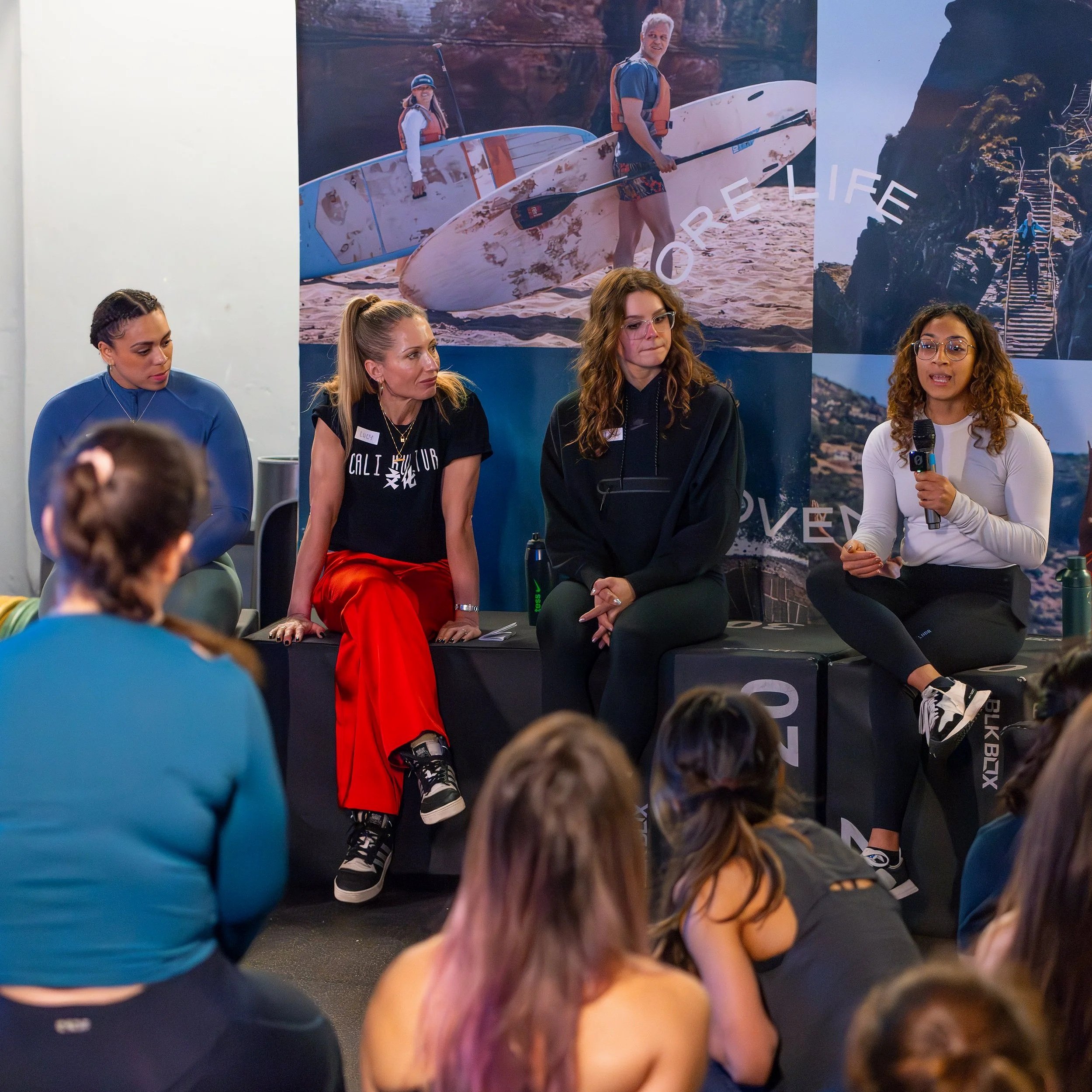 Four women sitting on a panel at a speaker event, with an audience seated in front of them. Behind the panelists are large posters depicting outdoor adventure scenes, including paddleboarding and hiking.