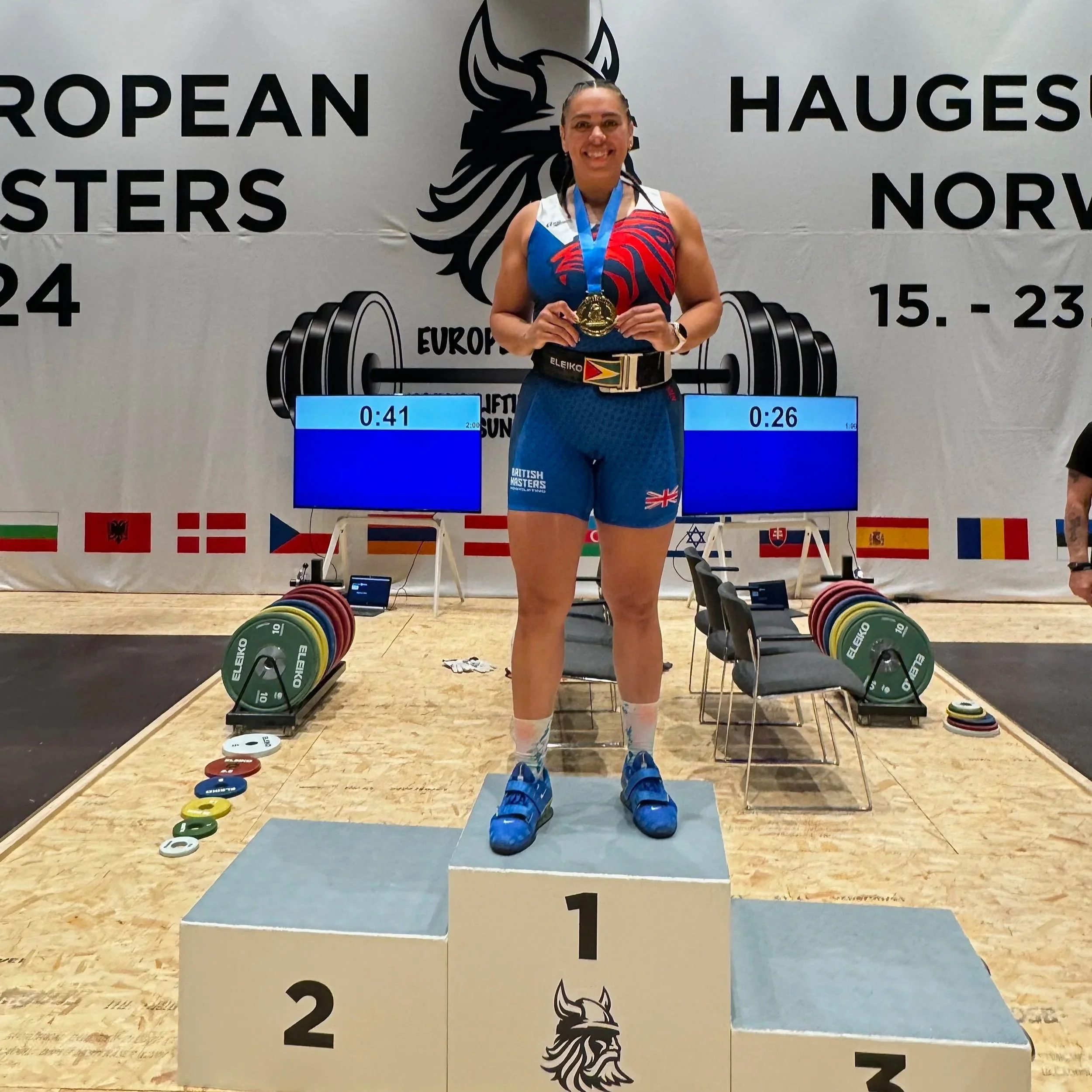 A female weightlifter standing on the first-place podium, holding a gold medal and smiling after her victory at a European weightlifting competition. She is dressed in a blue sports uniform with the Union Jack. In the background, there are computers, weight plates, and a banner with flags and the event details.