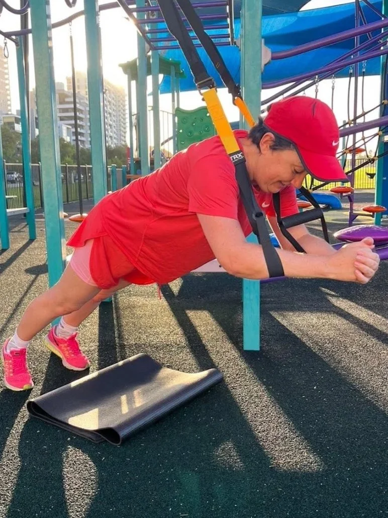 A woman in a red baseball cap, red Nike T-shirt, and pink sneakers doing a plank exercise on an outdoor playground surface. She is holding a forearm plank position with a black exercise mat on the ground beneath her, with playground equipment and city buildings in the background.